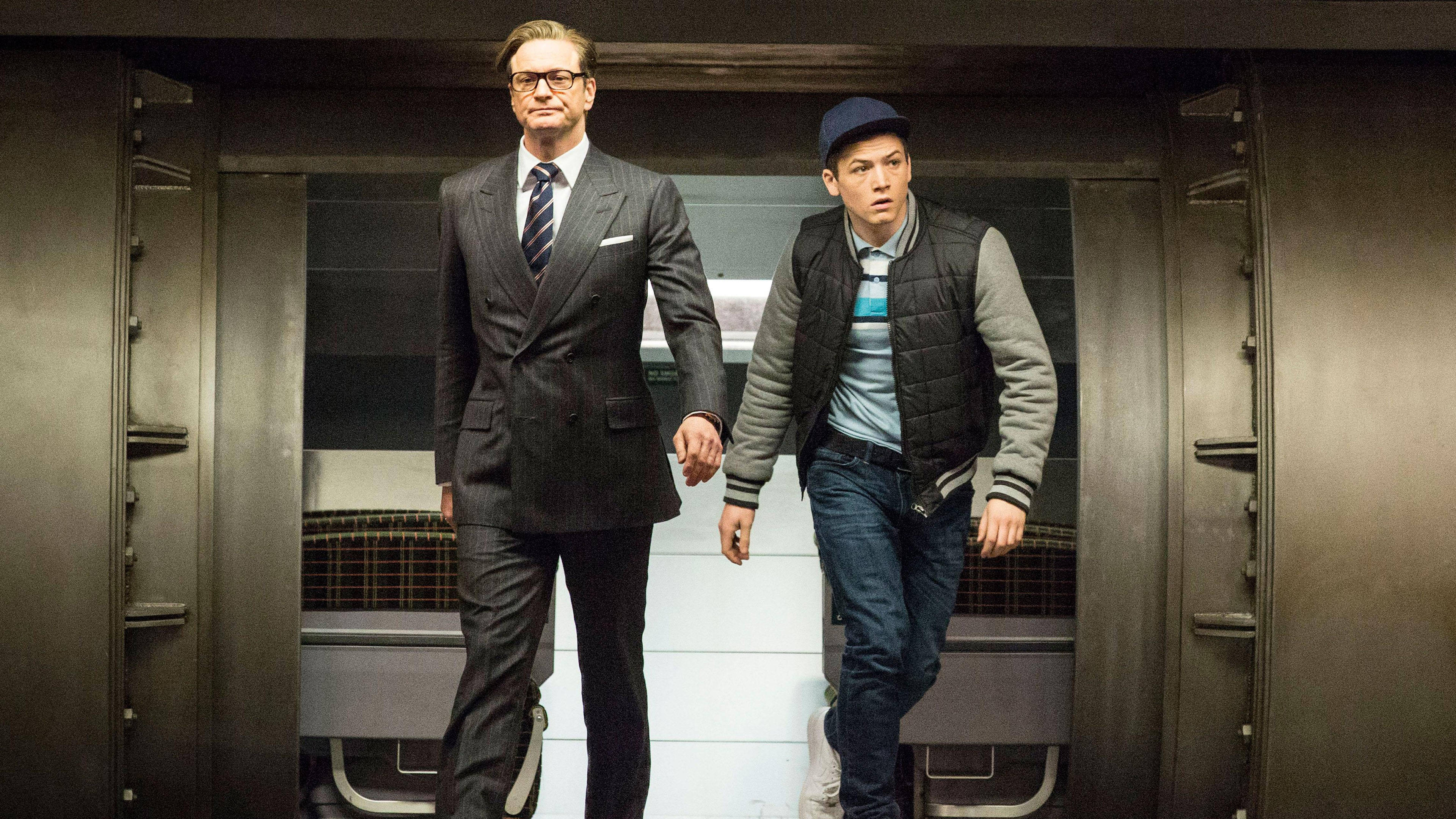 《王牌特工》Eggsy 與 Galahad 動作場景 壁紙 — 兩個男人手拖手穿過金屬走廊，一個穿西裝，另一個穿背心和帽子。
