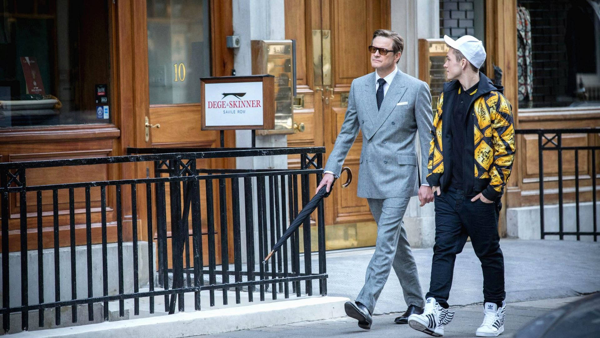 Eggsy 與加拉哈德 金士曼 壁紙 — 兩個男人在城市行人道上漫步，一個穿著俐落的灰色西裝拿著雨傘，另一個穿著大膽的黃色外套和白色帽子。