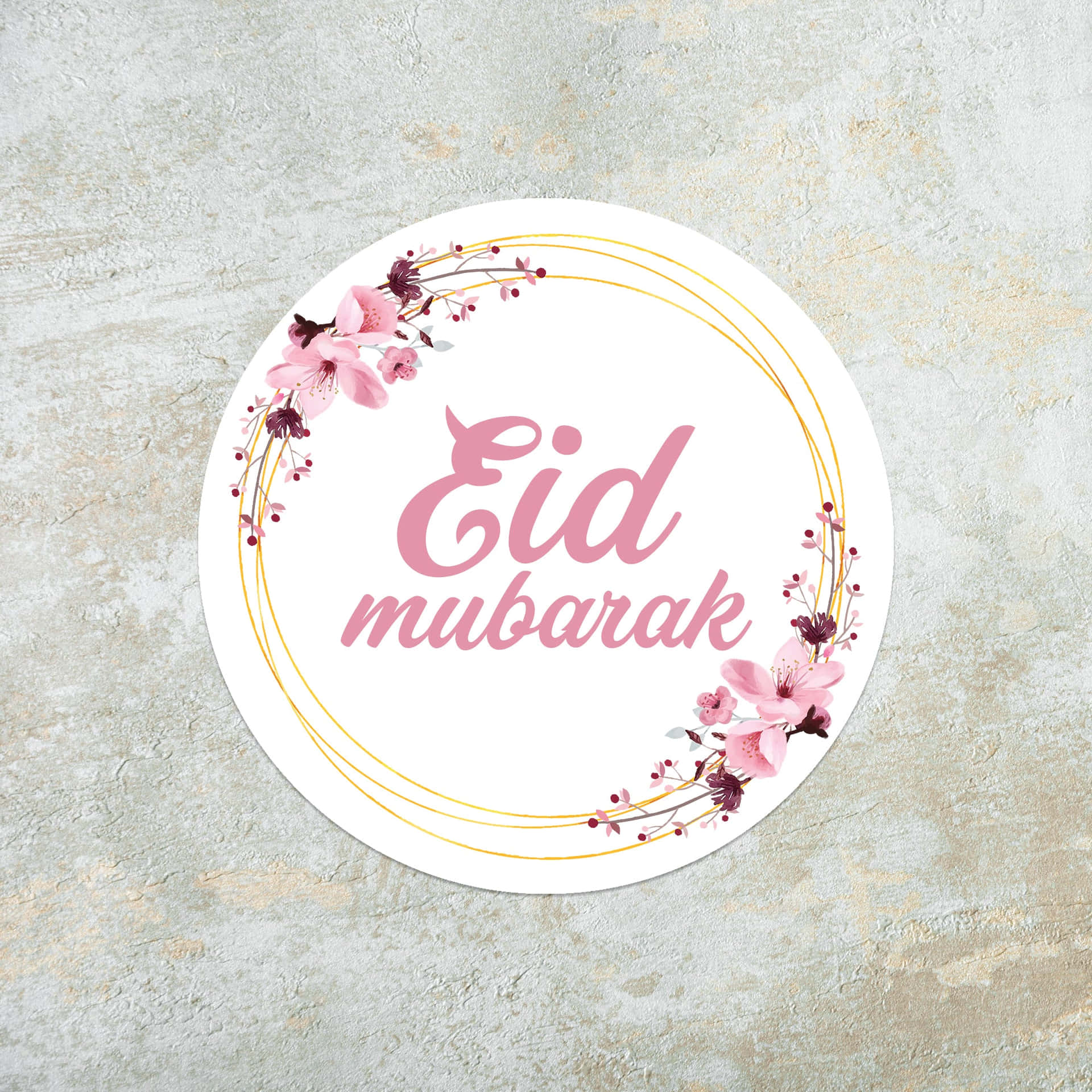 圓形 Eid mubarak 問候貼紙，帶有粉紅色花朵和金色線條，位於紋理灰色表面上。