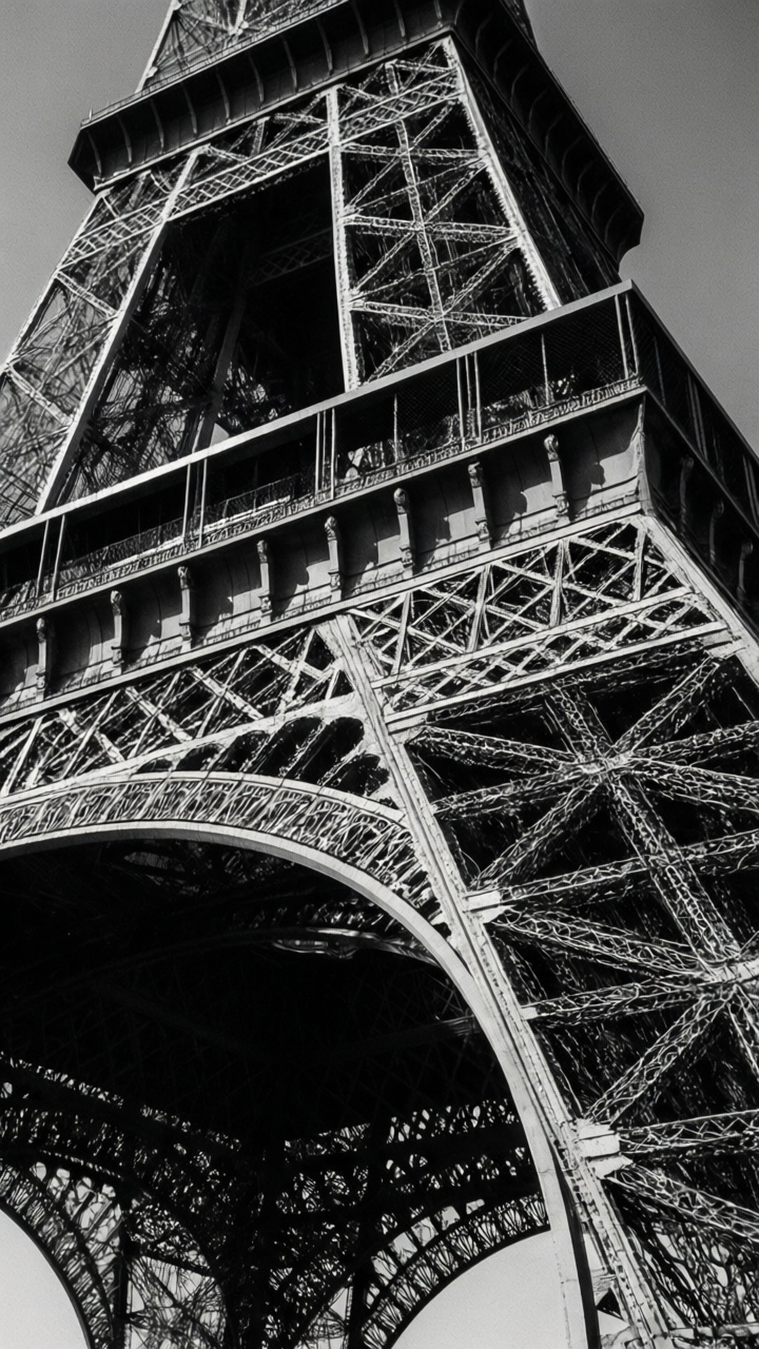 Eiffel in Monochrome 壁紙 — 一張從低角度拍攝的戲劇性黑白照片，捕捉了艾菲爾鐵塔底部複雜的鐵格柵和宏偉的拱門。