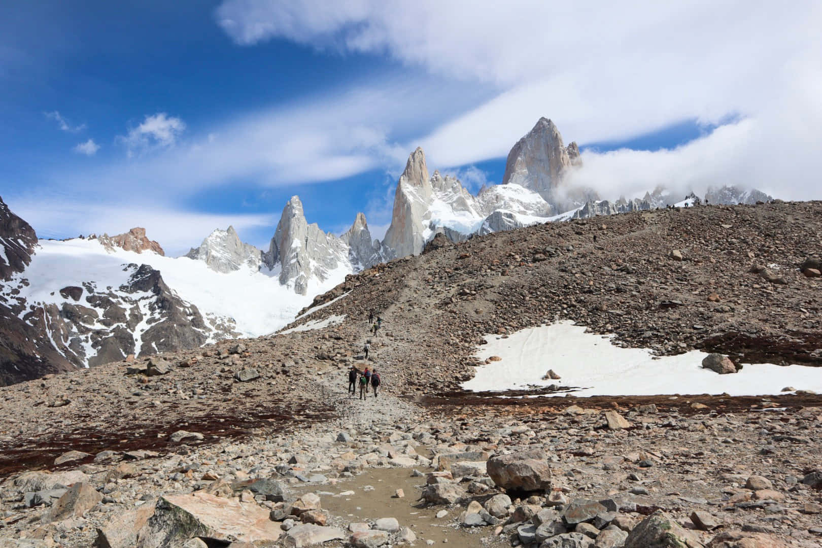 El Chalten的雪山風景 壁紙 — 遠足者沿著岩石高山小徑，在明亮藍天和薄雲下，向著積雪的山峰前進。