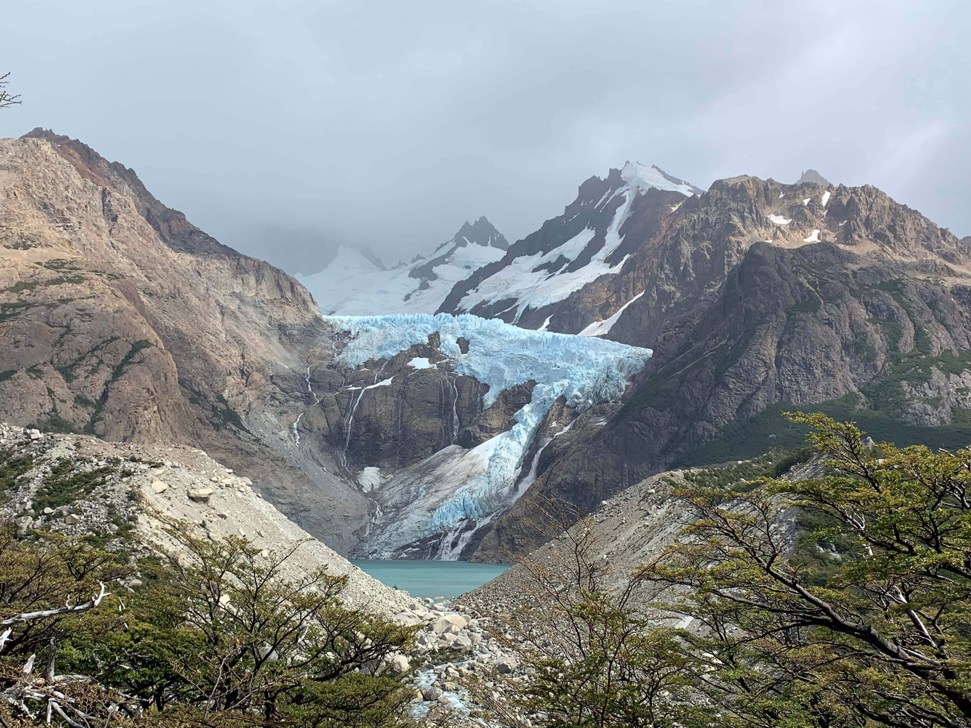 El Chalten的壯麗山景 壁紙 — 宏偉的冰川從崎嶇的山脈傾瀉而下，流入一個被深綠色樹木環繞的綠松石色湖泊，天空多雲。
