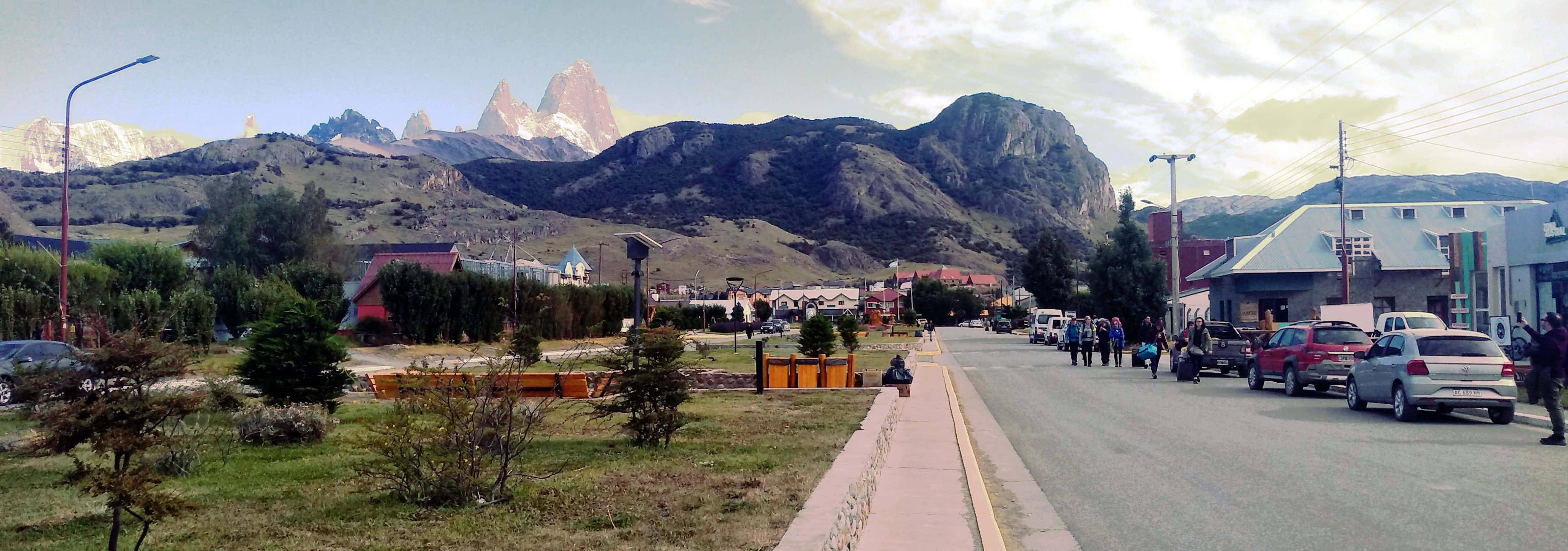 El Chalten小山村的風景照 壁紙 — 一條小鎮街道，停泊的汽車和行人，背景是部分多雲的天空下高聳的雪山。