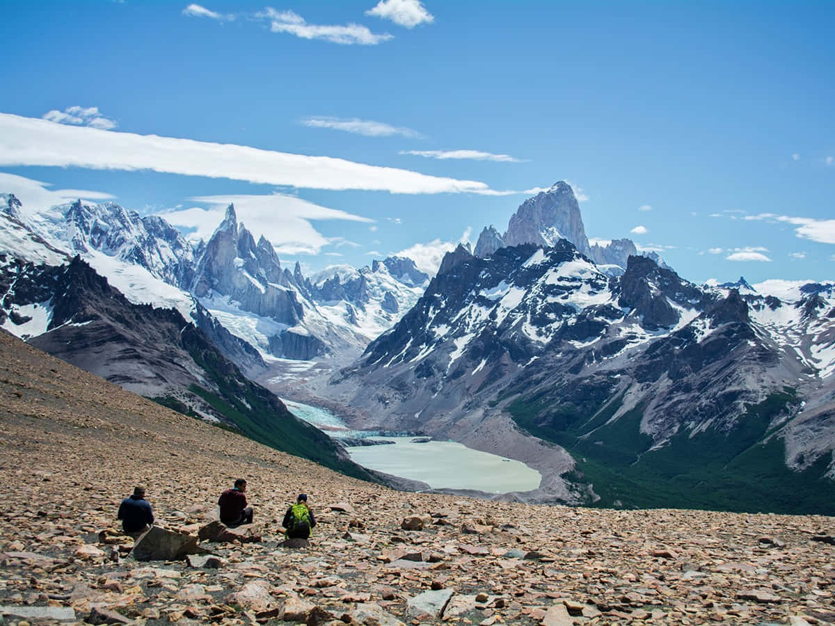 El Chalten令人驚嘆的山景 壁紙 — 三名遠足者在岩石斜坡上休息，俯瞰冰川湖和崎嶇的雪峰，頭頂是明亮的藍天。
