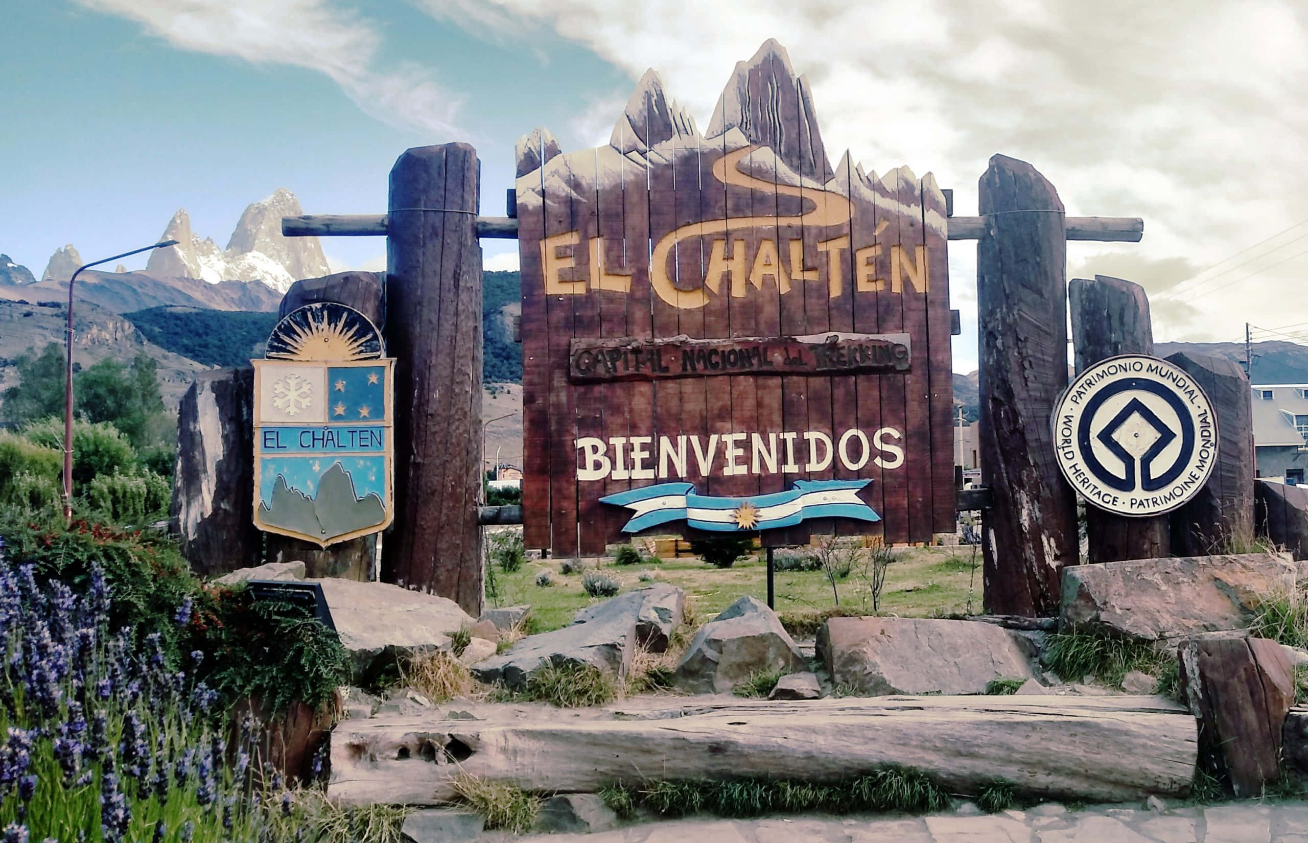 El Chalten山嶺天堂的大型迎賓標誌 壁紙 — 一個質樸的木製歡迎牌，為埃爾查爾滕而設，矗立在部分多雲的天空下，鋸齒狀的雪山前。