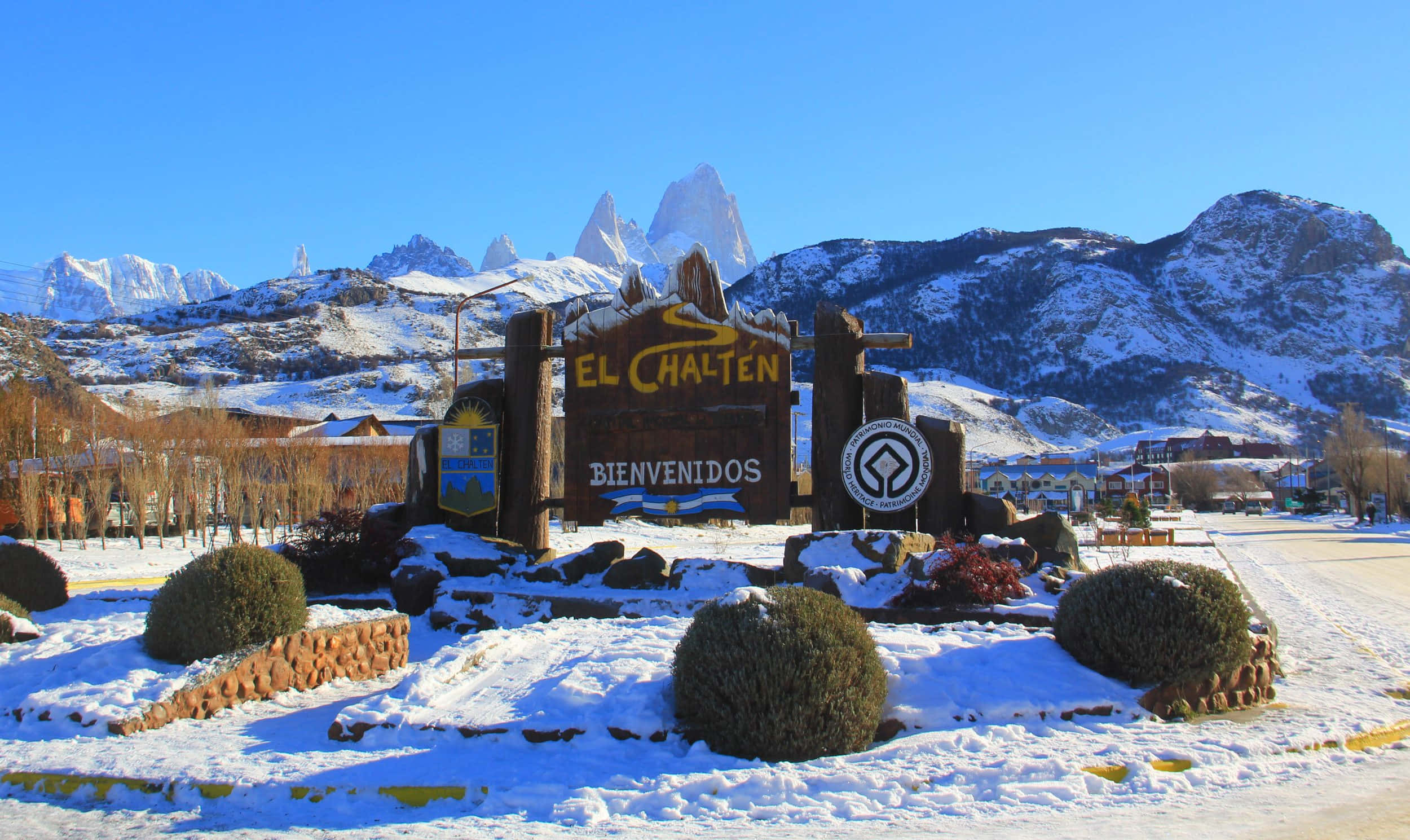 El Chalten山嶺村的冰原風景 壁紙 — 一個積雪的埃爾查爾滕歡迎標誌，矗立在晴朗藍天下雄偉的雪山前。