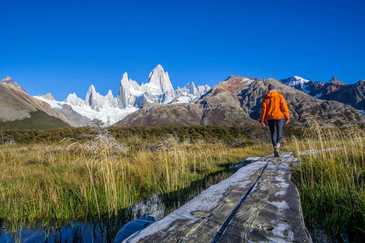El Chalten村莊的愜意一日 壁紙 — 一名身穿橙色外套的遠足者沿著木棧道穿過草地濕地，走向晴朗藍天下雄偉的菲茨羅伊雪山峰頂。