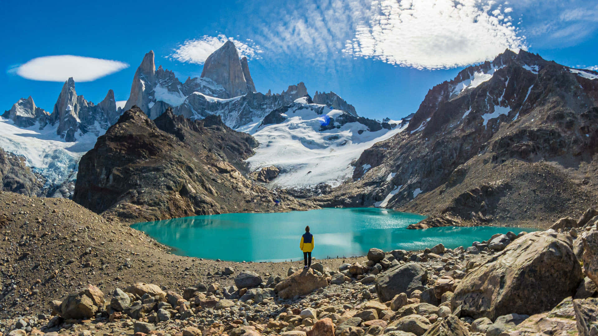 El Chalten的全景湖景 壁紙 — 一位身穿黃色外套的孤獨遠足者站在岩石地形上，面前是充滿活力的綠松石色湖泊，周圍是白雪覆蓋的鋸齒狀山峰，天空湛藍，雲朵輕柔。