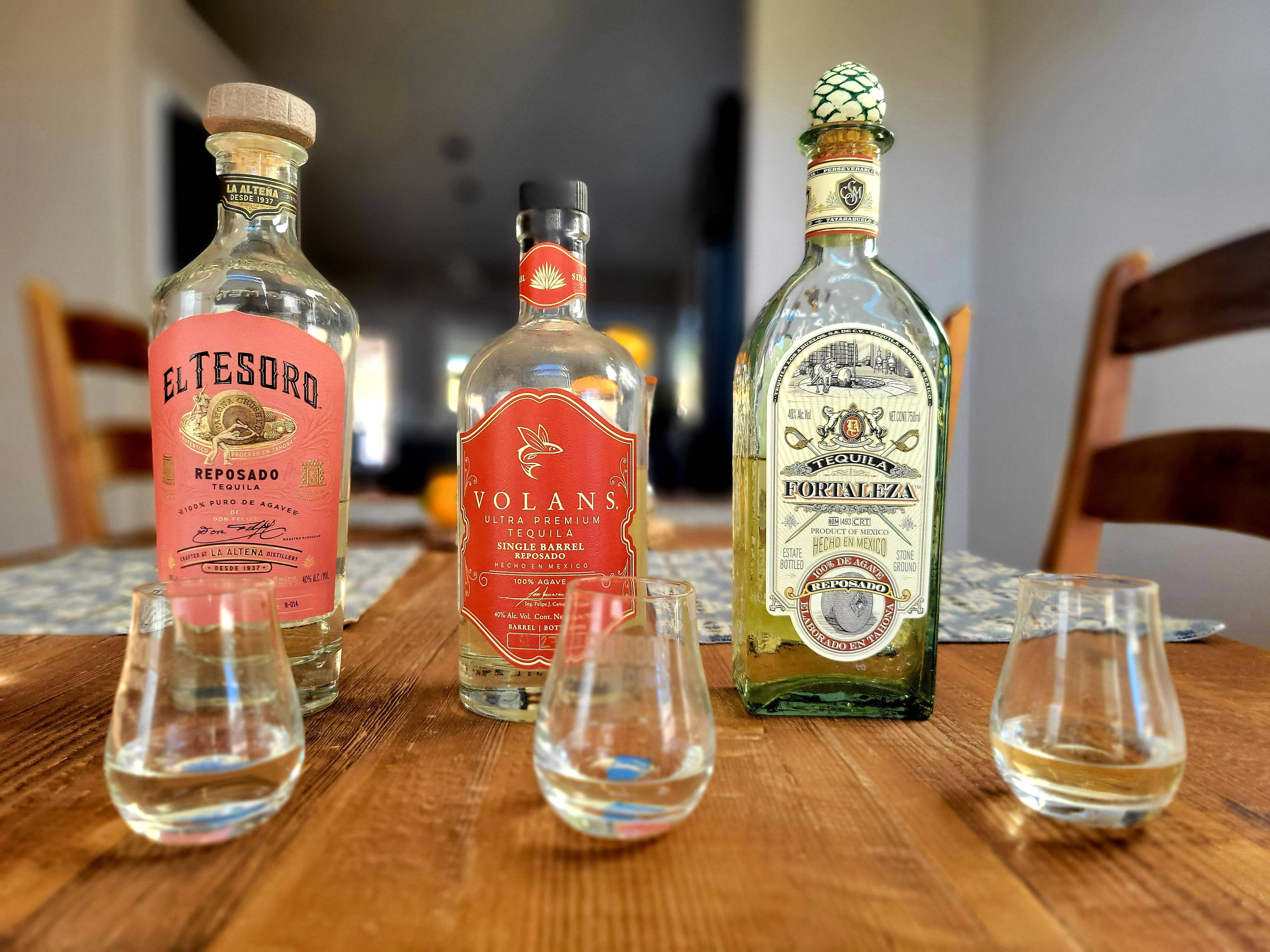 El Tesoro、Volans 與 Fortaleza 龍舌蘭酒 壁紙 — 三種不同的龍舌蘭酒瓶配品酒杯，擺放在一張鄉村風格的木桌上，室內燈光溫暖。