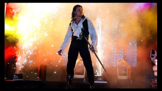 El Zorro 優雅登場 壁紙 — 一位穿著海盜服裝的表演者站在煙霧瀰漫的舞台上，手持劍，周圍火花四濺，在彩色燈光下。