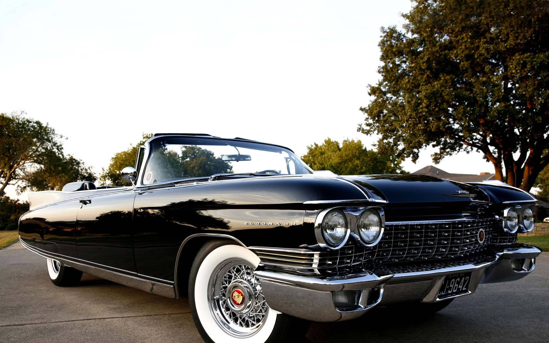 黑色 Eldorado Biarritz 凱迪拉克 iPhone 版 壁紙 — 一架閃亮黑色1960年代凱迪拉克開篷車在柔和日光下閃耀，停泊在綠樹成蔭的住宅街道上。