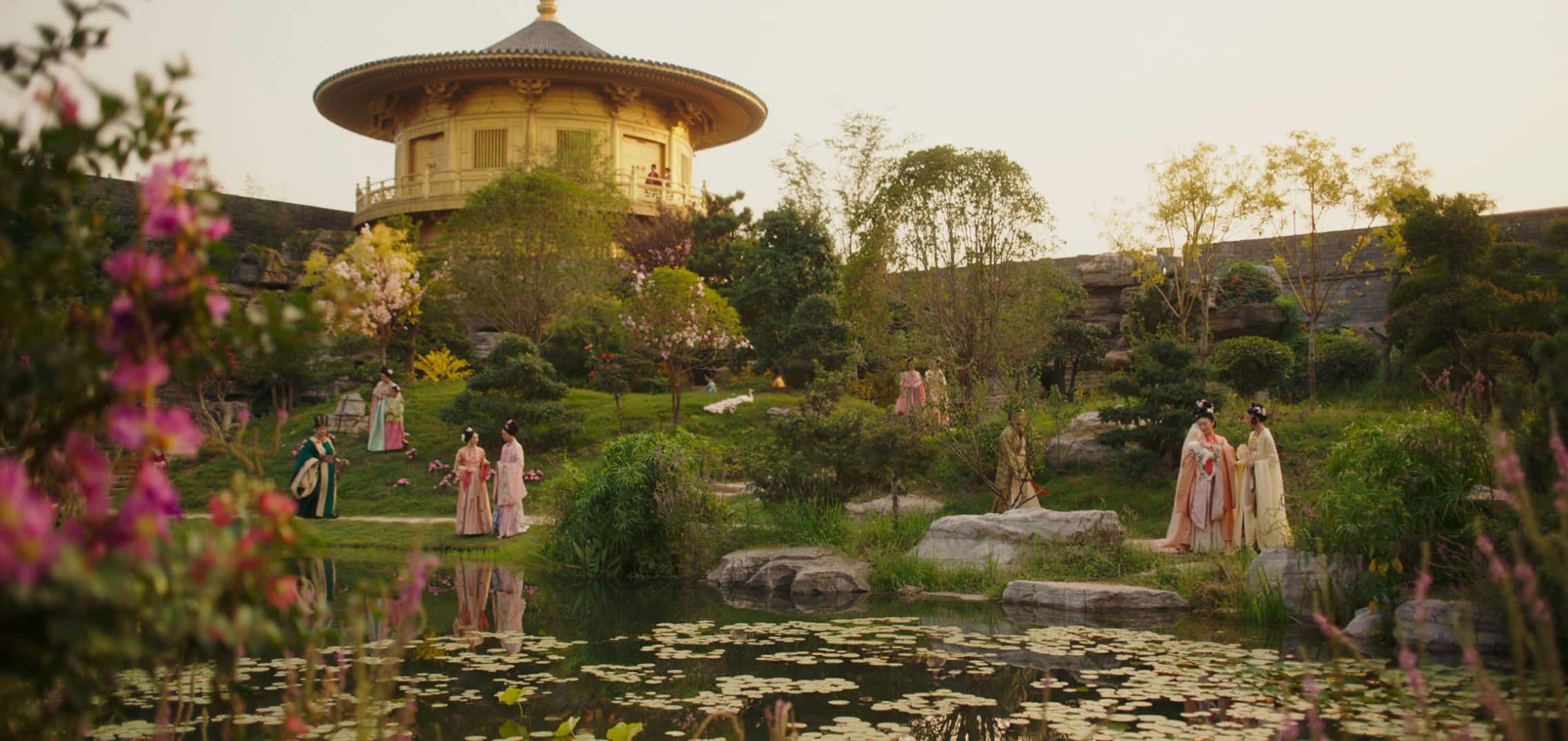 穿著傳統長袍的人們走過一個有金色寶塔和百合池塘的茂盛花園。