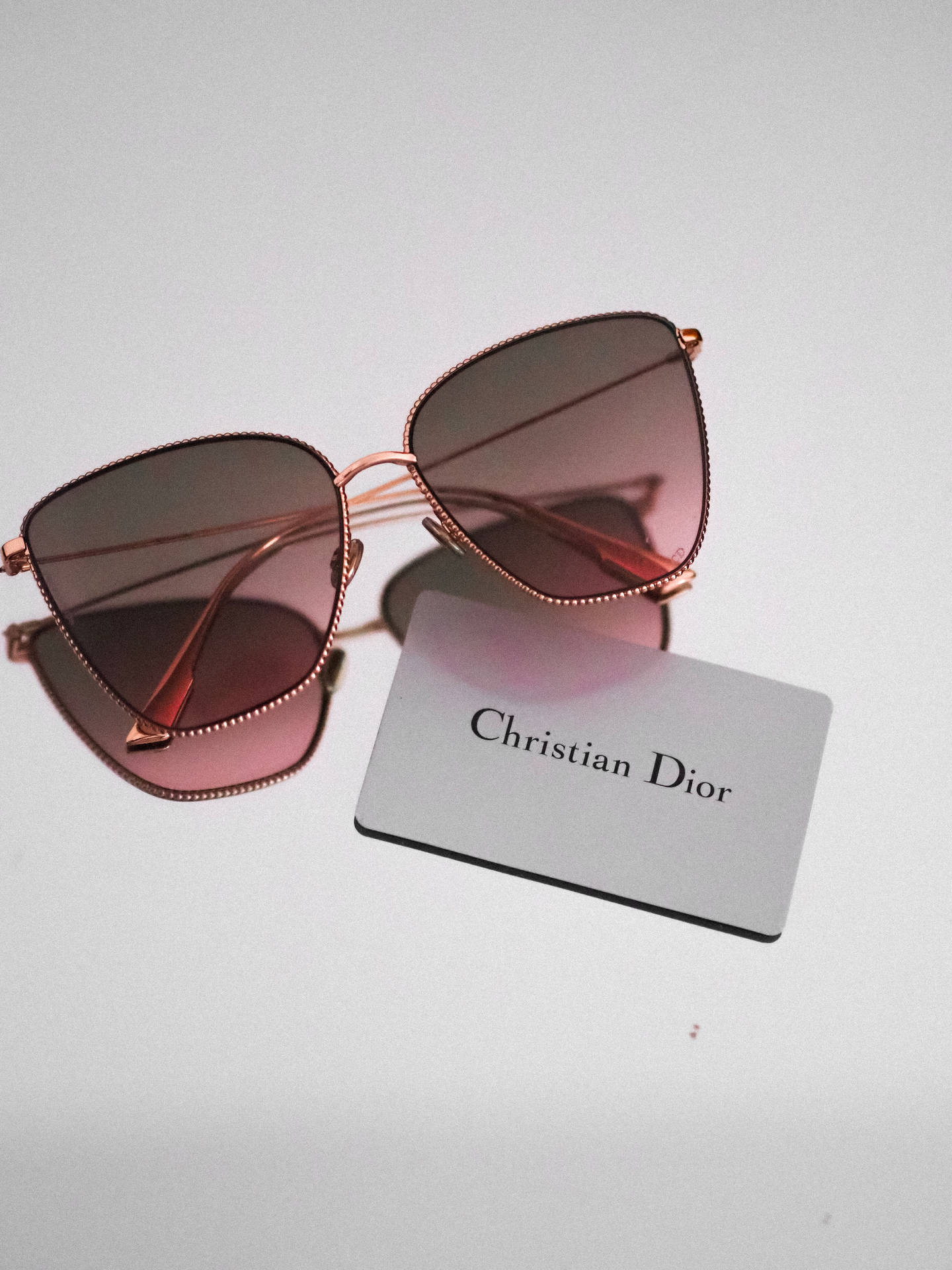 Christian Dior太陽眼鏡 壁紙 — 優雅的玫瑰金色Christian Dior太陽眼鏡，配以漸變粉紅色鏡片，放在簡約白色表面上，旁邊有一張品牌卡。