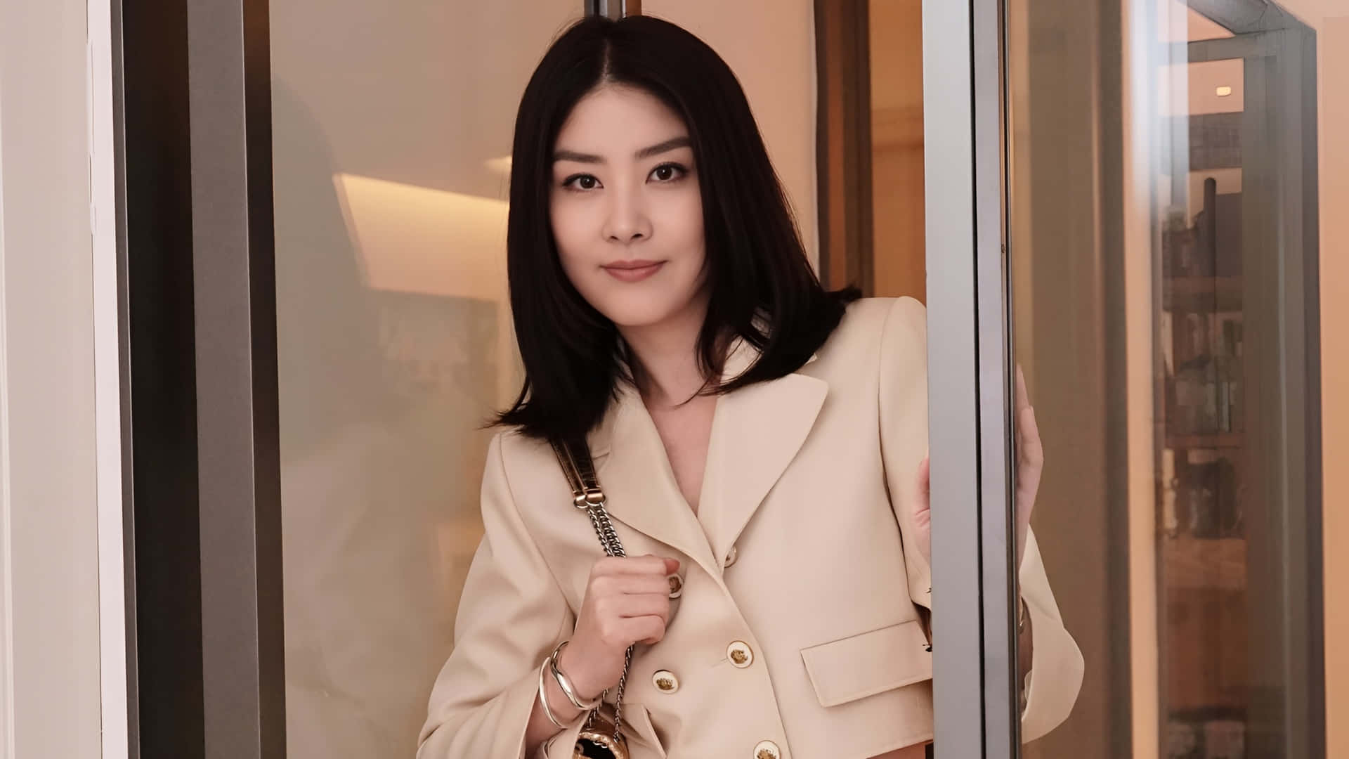  壁紙 — 一位身穿米色西裝外套的優雅女士，站在現代玻璃門前，露出溫柔的微笑。