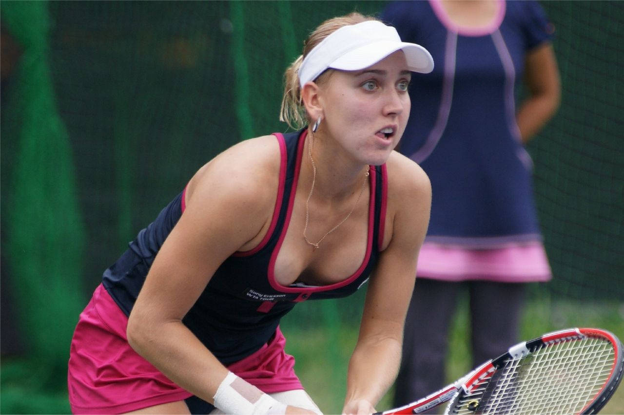 Elena Vesnina 預備擊球姿勢 壁紙 — 一位專注的女網球運動員戴著白色遮陽帽和粉紅色裙子，蹲在綠色球場上準備回擊。