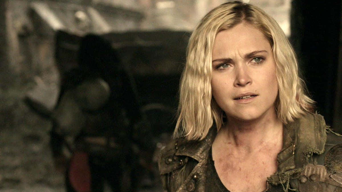 Eliza Taylor 焦慮神情特寫 壁紙 — 一位金髮女子穿著破舊的夾克，在昏暗骯髒的環境中，帶著緊張的表情專注地凝視。
