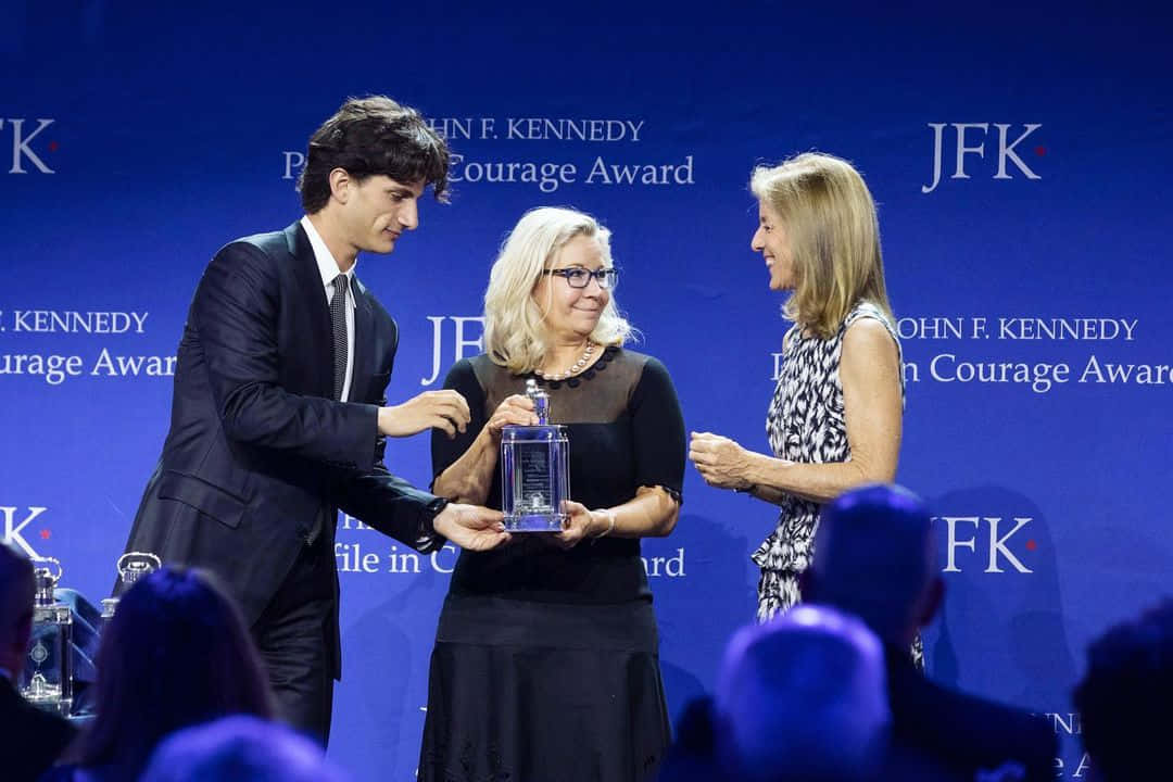 Elizabeth Cheney 領取獎項 壁紙 — 一位身穿西裝的年輕男子在藍色舞台背景上，帶有JFK Courage Award文字，向一位年長女性頒發玻璃獎項。