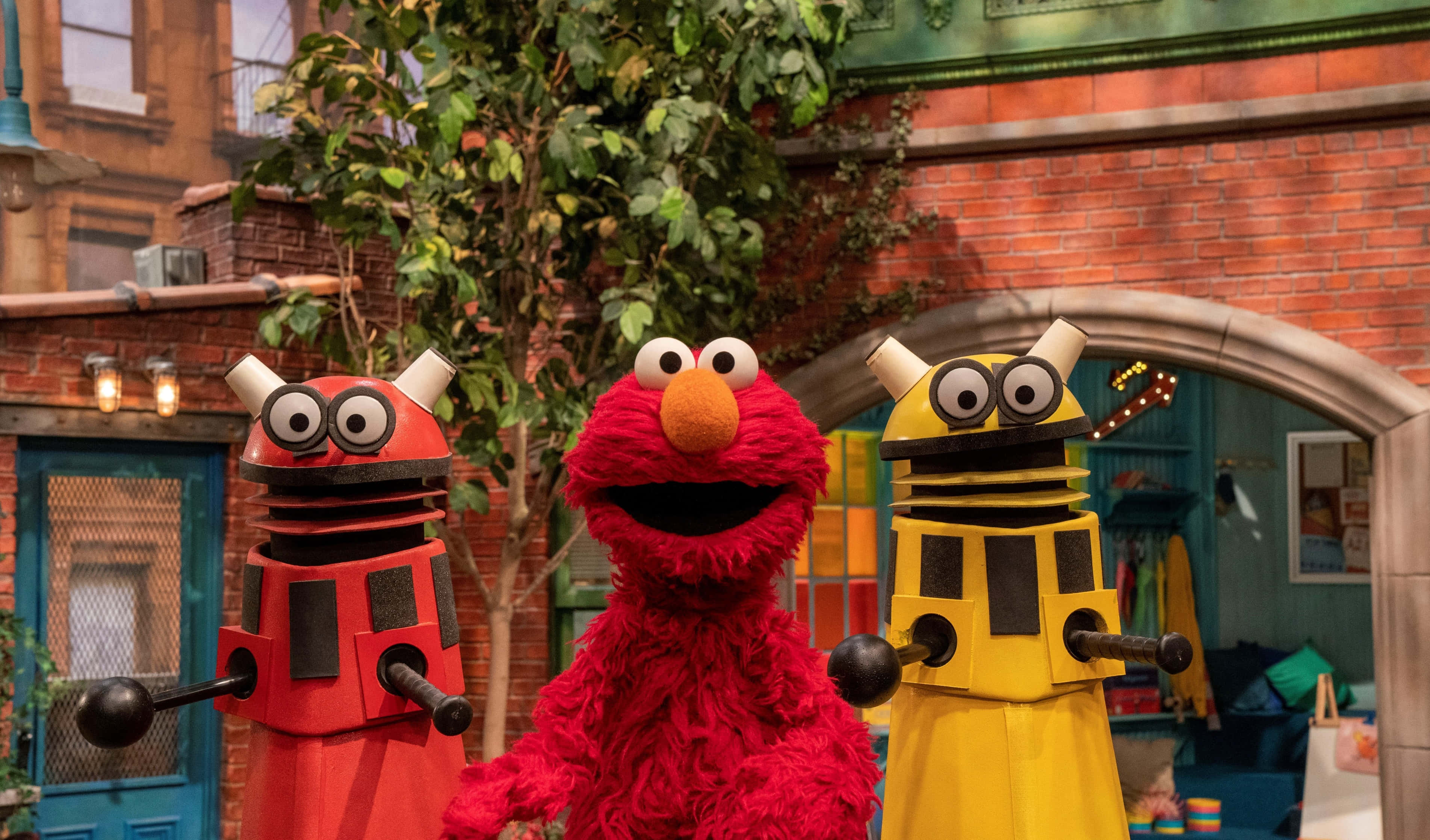 Elmo 桌布 壁紙 — Elmo 愉快地站在兩個色彩繽紛的機械人公仔之間，背景是充滿活力的芝麻街庭院，有磚砌建築和綠植。