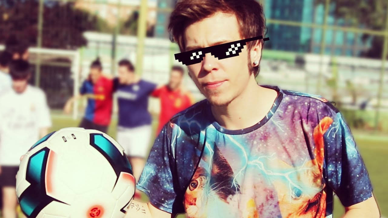 elrubiusOMG 穿著經典匪幫風格 壁紙 — 一個年輕男子戴着像素化太陽眼鏡，穿着銀河貓T恤，在陽光明媚的球場上拿着足球，背景有隊友。