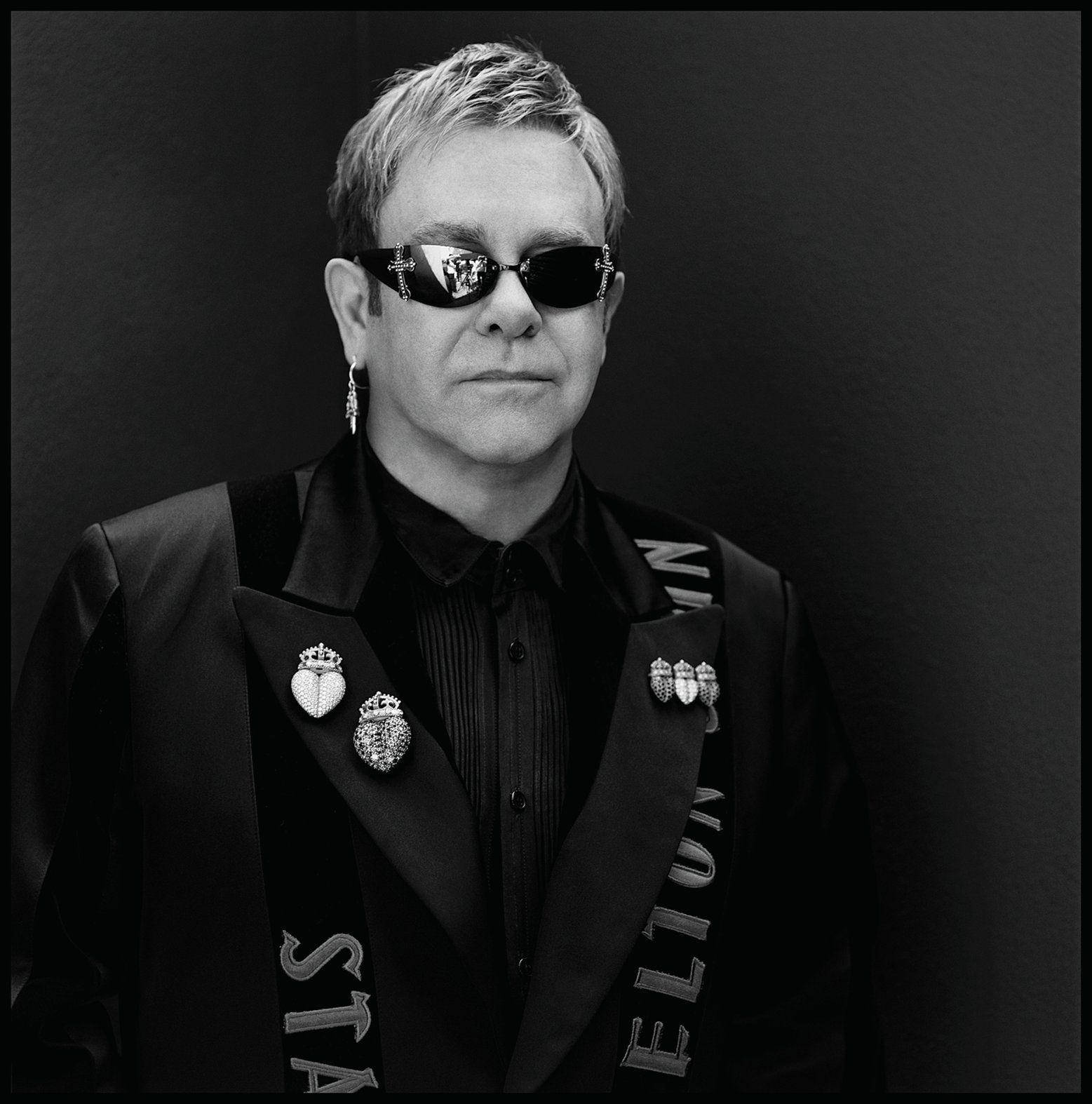 Elton John 黑色 Rocketman 壁紙 — 艾爾頓·約翰身穿黑色燕尾服，佩戴裝飾別針和太陽眼鏡，在深色背景前擺姿勢。