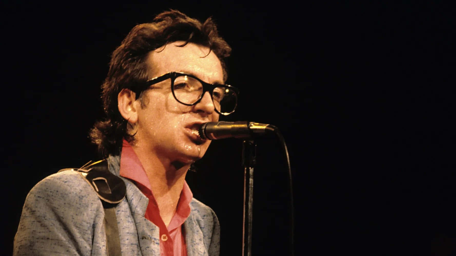 Elvis Costello 1979年經典影像 壁紙 — 一個戴著粗黑框眼鏡的男人，在黑暗背景的舞台燈光下，對著麥克風深情演唱。