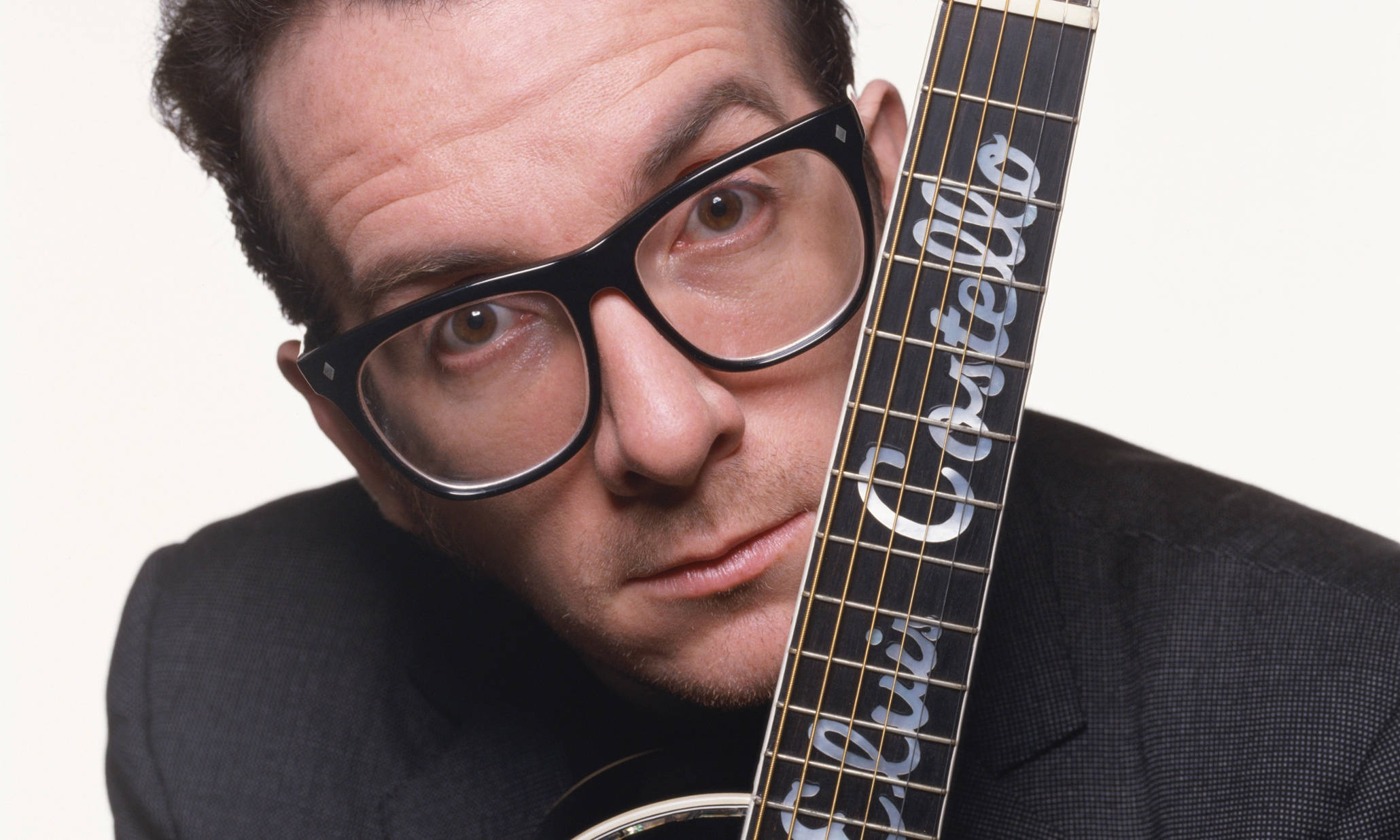 受愛戴的藝人Elvis Costello 壁紙 — 一個戴著粗黑框眼鏡的男人，從標有Elvis Costello的吉他琴頸後面，對著純白色背景凝視。