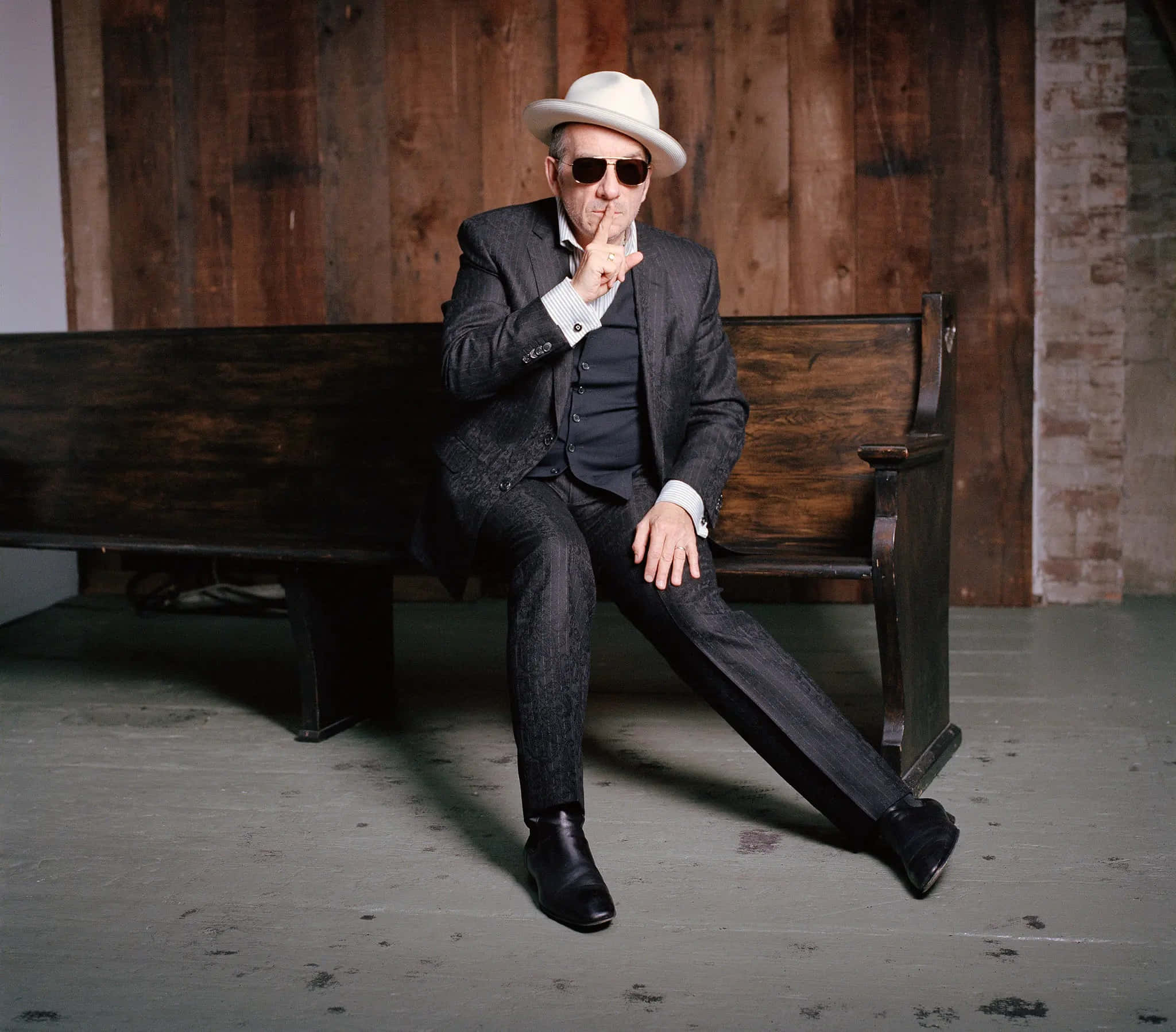 Elvis Costello 為《紐約時報》專訪拍攝 壁紙 — 一位穿著灰色西裝、白色軟呢帽和太陽眼鏡的時尚男士坐在木長椅上，做出噤聲手勢。