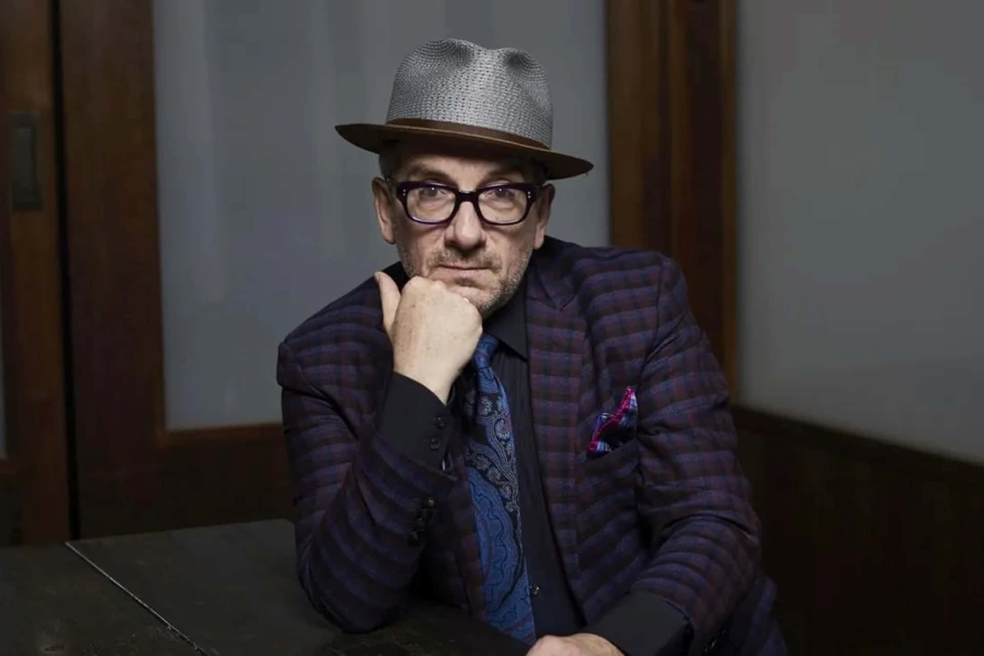 Elvis Costello 為《多倫多星報》專訪拍攝 壁紙 — 一個穿著圖案西裝和軟呢帽的男人，托著下巴，表情若有所思。