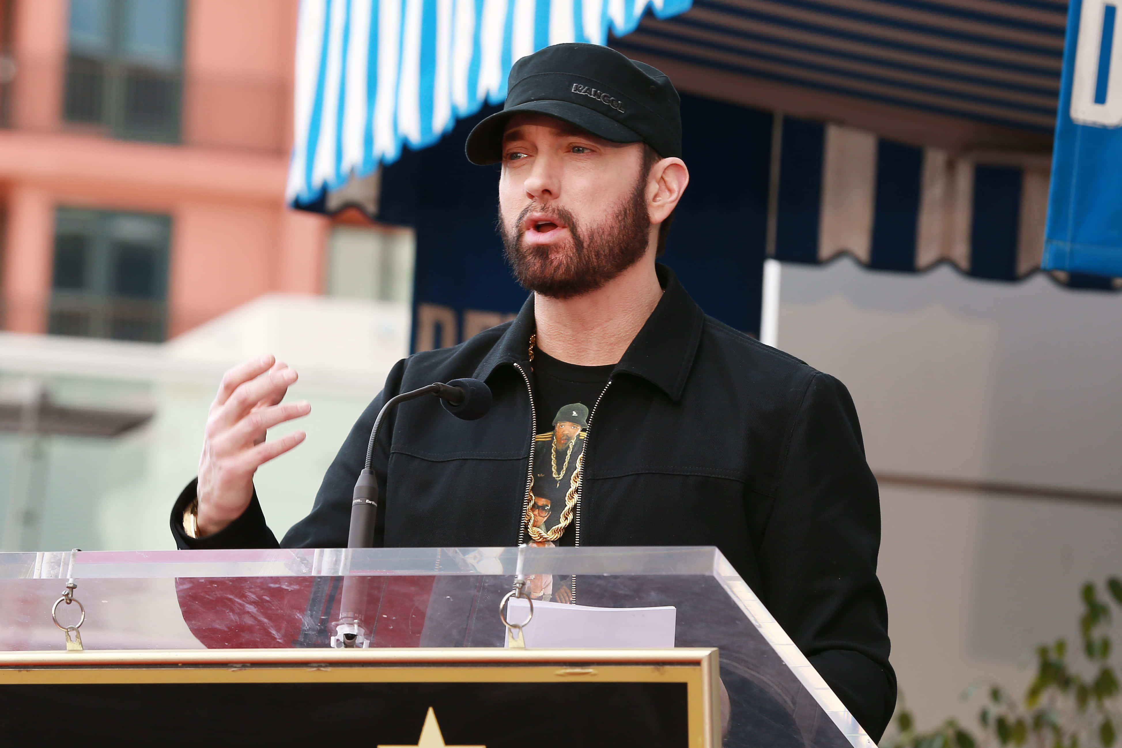 Eminem 桌布 壁紙 — Emil eminem 喺戶外講台上熱情演講，身後有條紋簷篷同建築物。