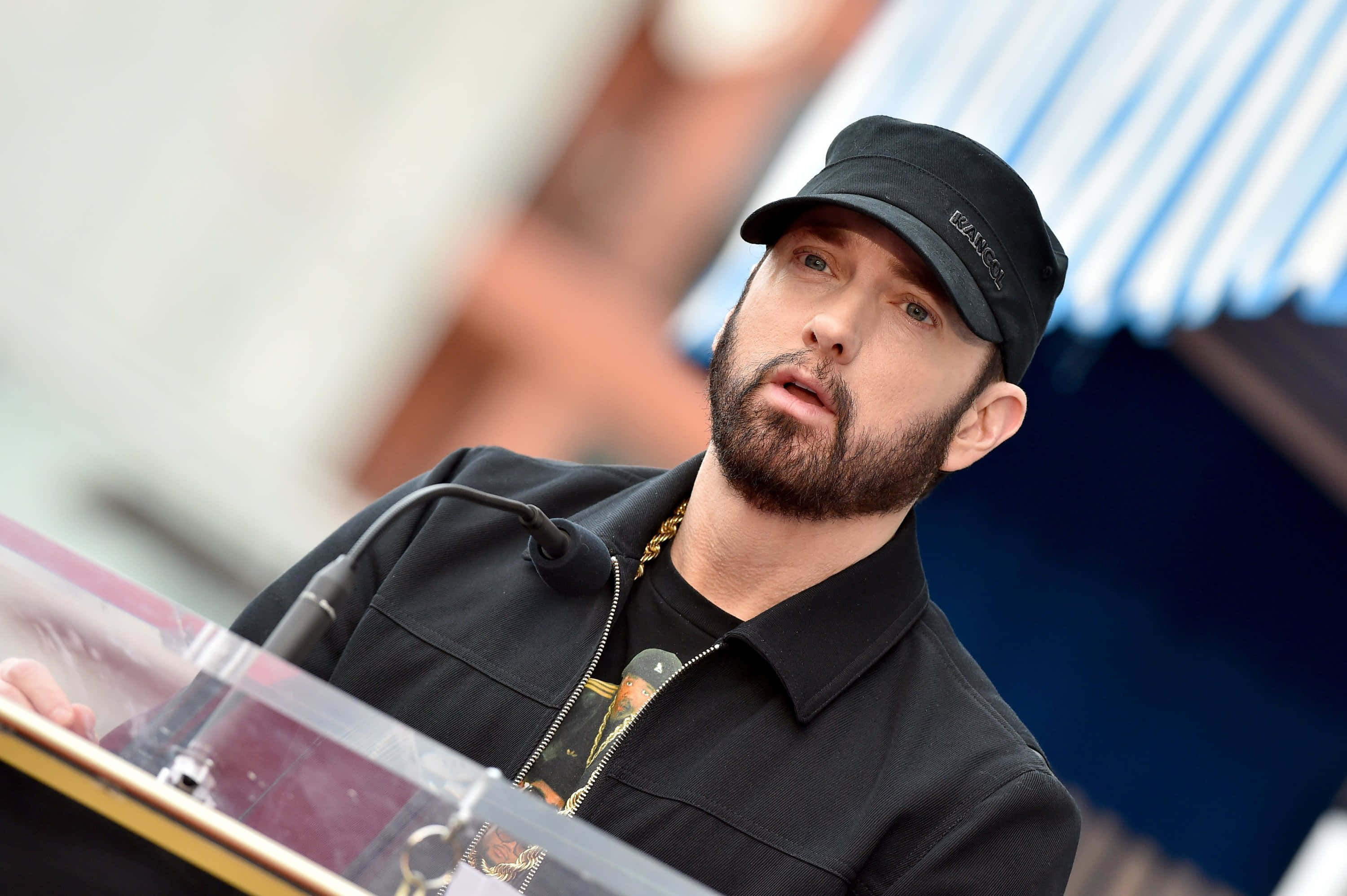 Eminem 桌布 壁紙 — Emil eminem 在講台上發言，戴著黑色帽子和外套，戴著金鏈，在明亮的日光下。