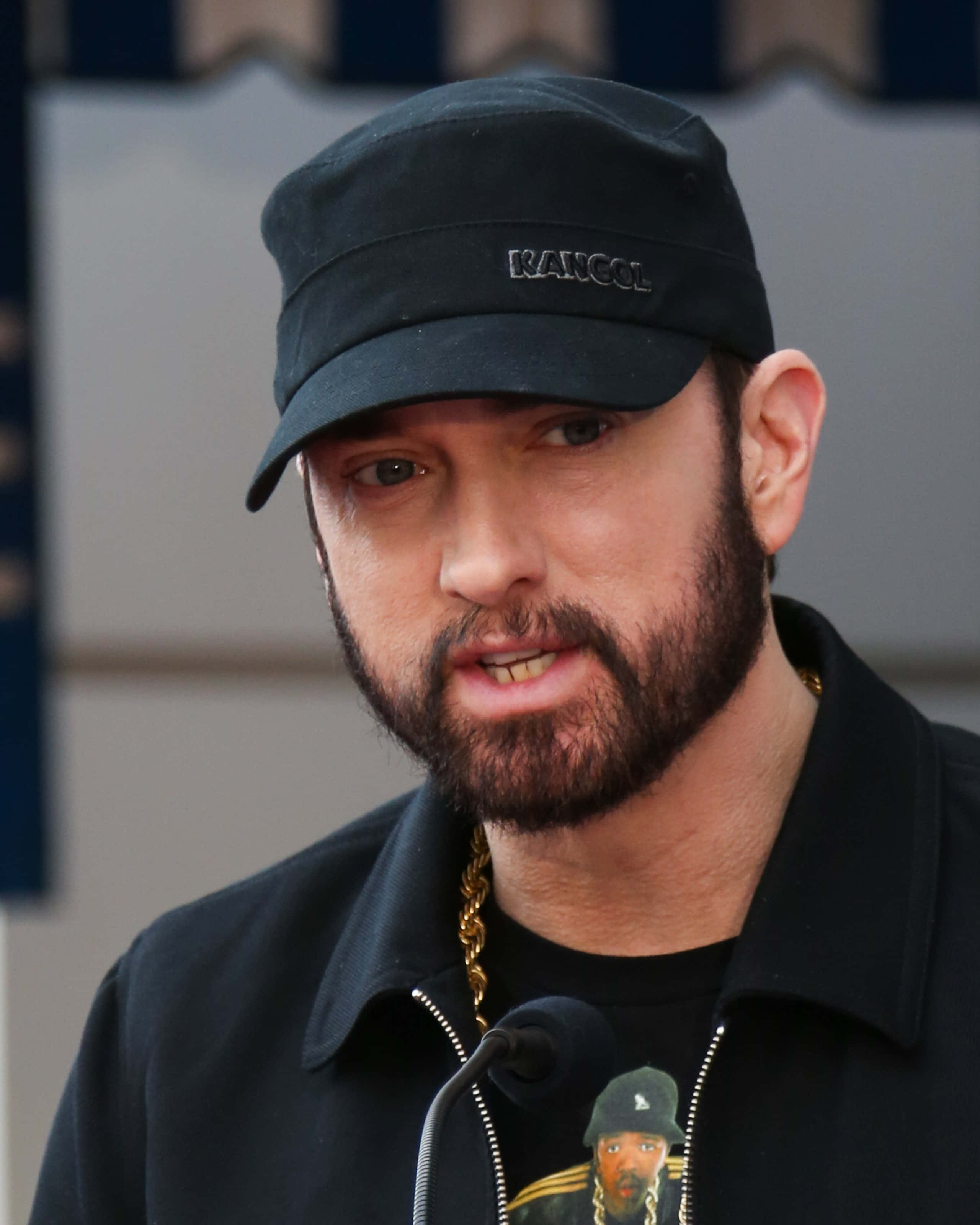 Eminem 桌布 壁紙 — Emil eminem 對著咪高峰說話，戴著黑色帽子和外套，配戴金鏈。
