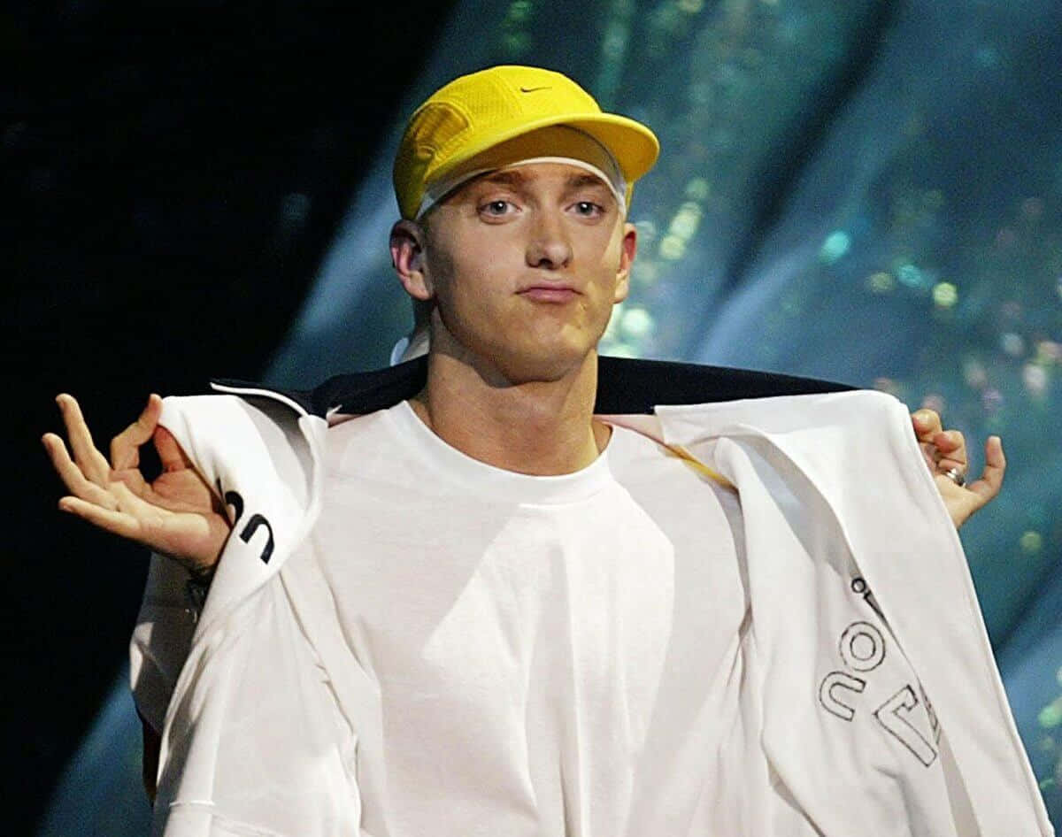 Eminem 桌布 壁紙 — Emil eminem 站在舞台上，戴著黃色帽子，穿著白色襯衫，在明亮的舞台燈光下將外套搭在肩上。