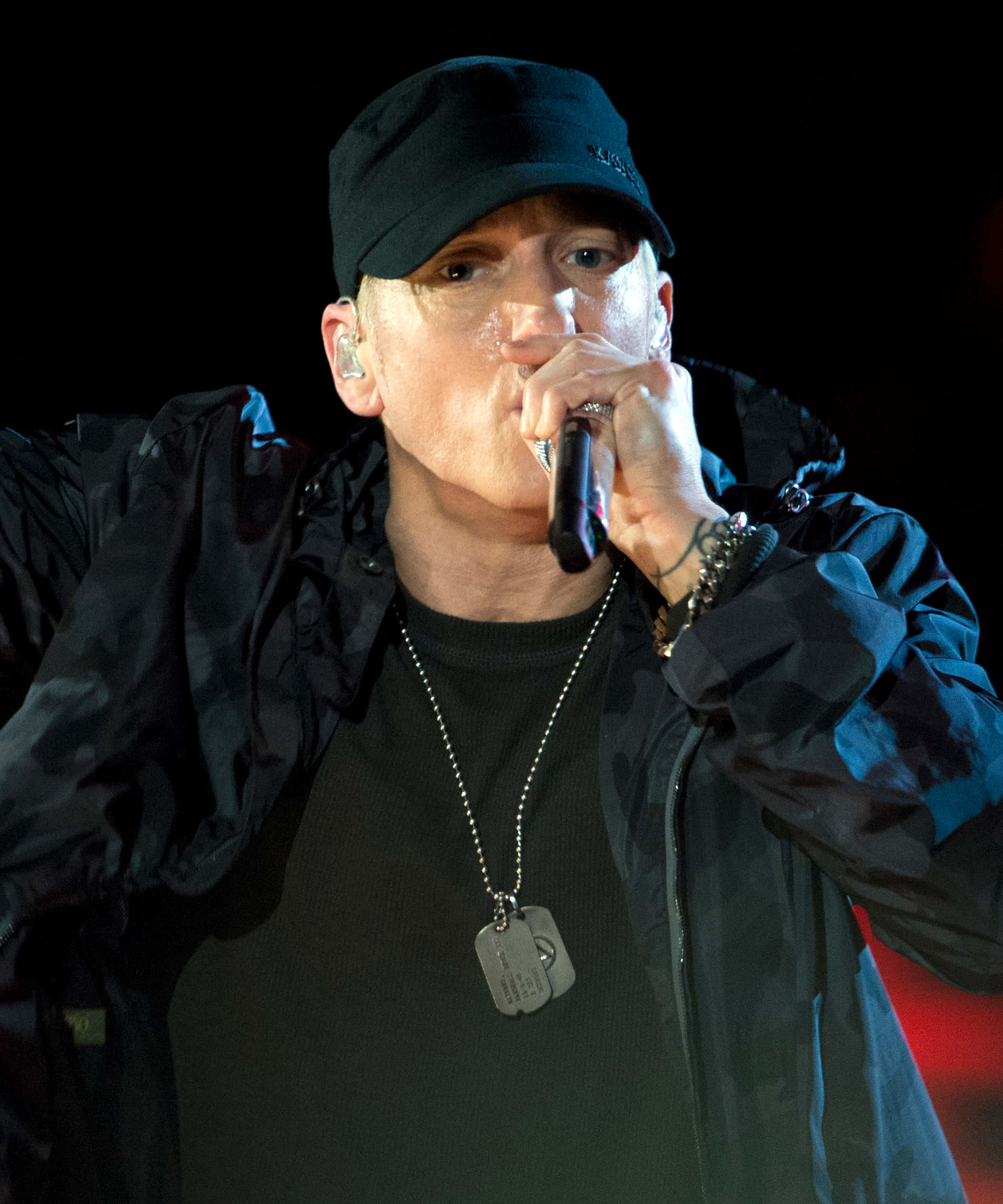 Eminem 桌布 壁紙 — Eminem 在舞台上表演，戴著黑色帽子和外套，在明亮的舞台燈光下將麥克風靠近嘴邊。
