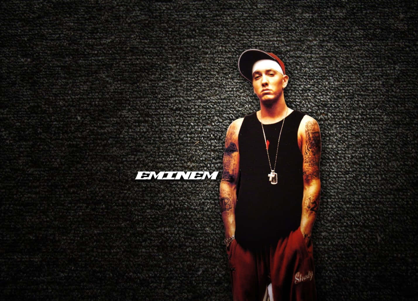 Eminem 桌布 壁紙 — Eminem 自信地站在深色紋理牆前，身穿黑色背心、紅色褲子和帽子，手臂上的紋身清晰可見。