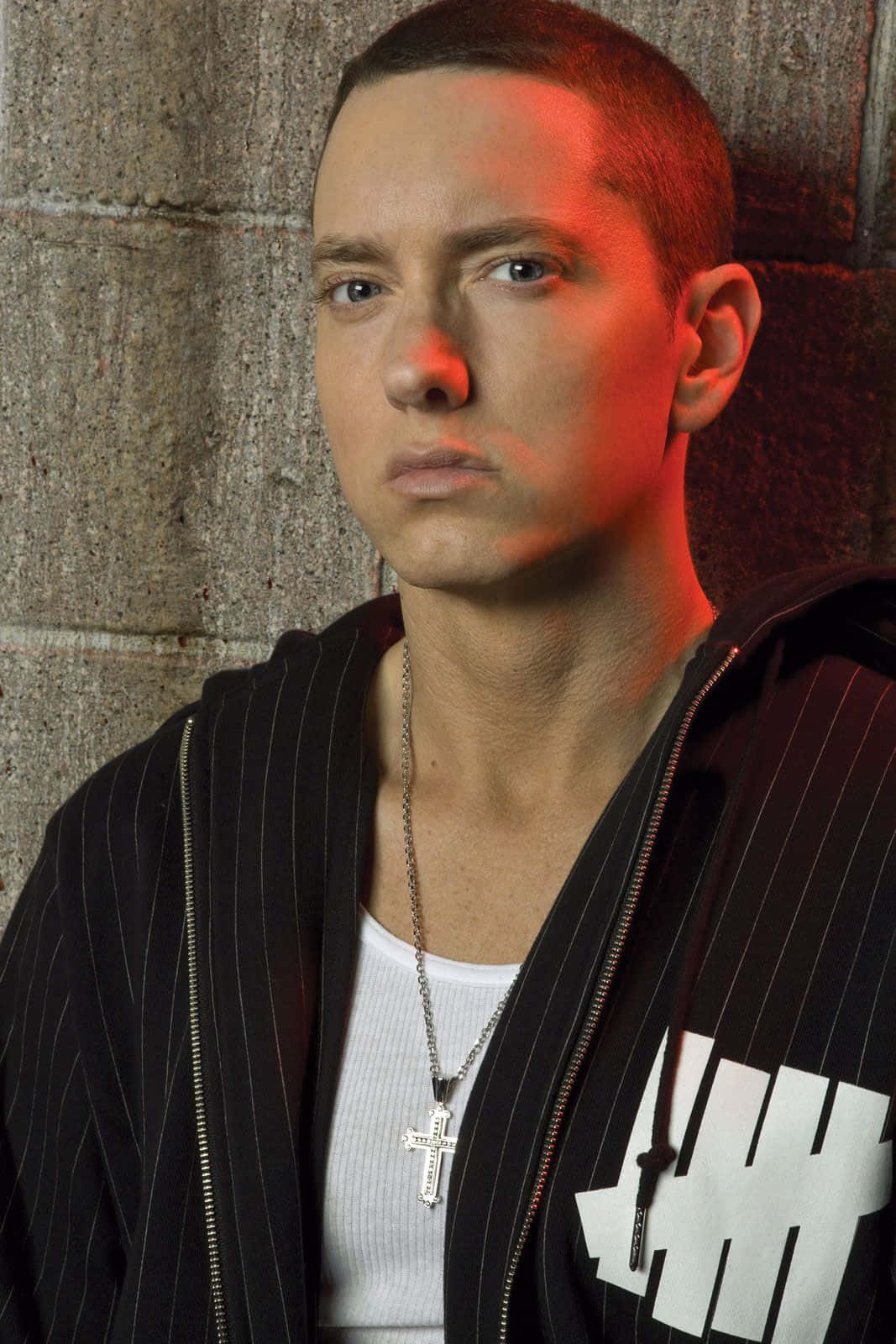 Eminem 桌布 壁紙 — Eminem 靠在混凝土牆上，在戲劇性的紅光下，穿著條紋連帽衫和十字架項鍊。