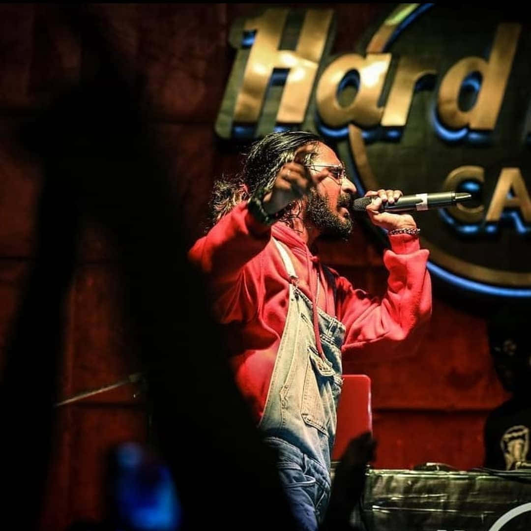 Emiway Bantai 在 Hard Rock Cafe 演出 壁紙 — 一位留鬍鬚的男子身穿紅色連帽衫和牛仔布工裝褲，在燈光昏暗的舞台上對著麥克風激情演唱，身後是發光的 Hard Rock Cafe 標誌。