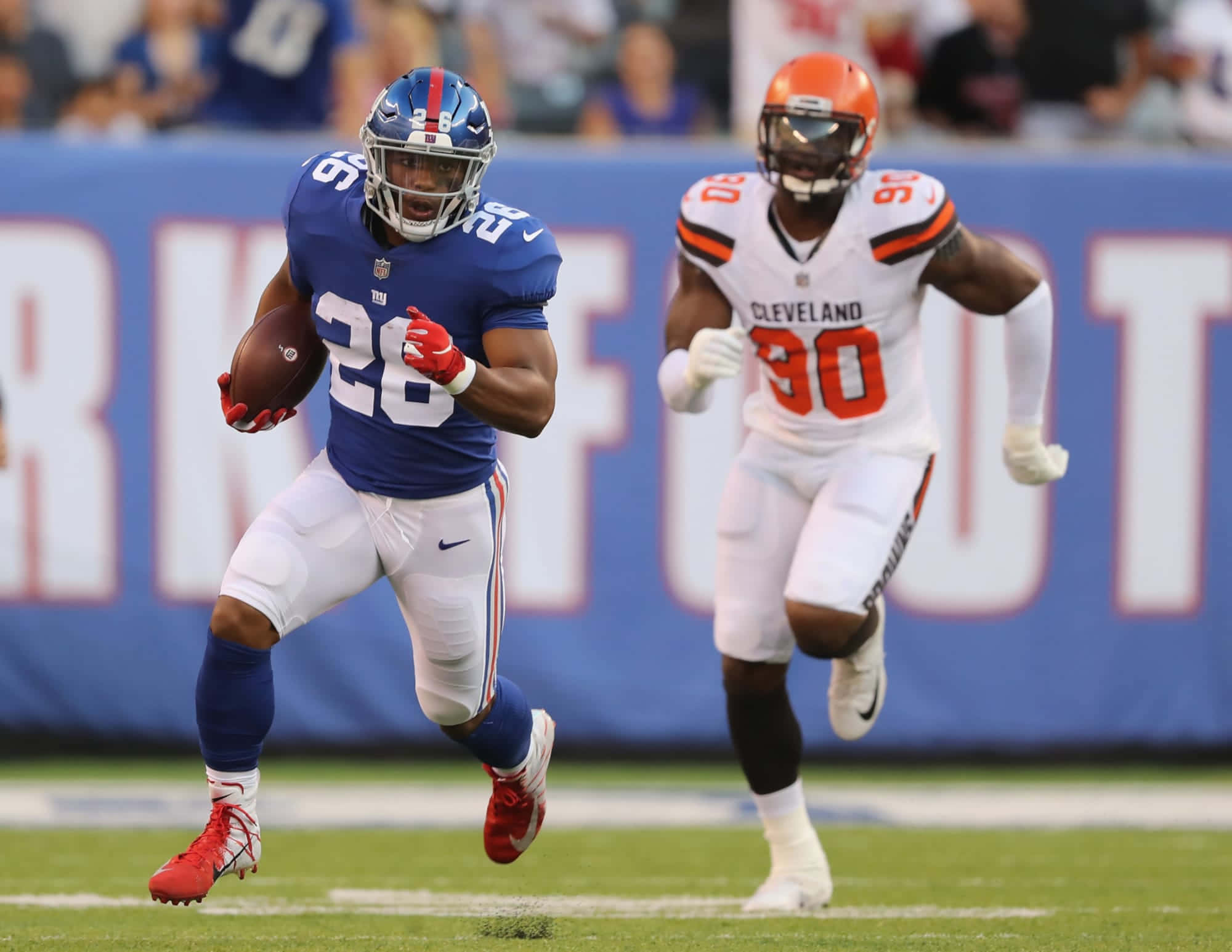 Emmanuel Ogbah 足球賽場風采 壁紙 — 紐約巨人隊的跑鋒身穿藍色球衣，帶著橄欖球衝刺，而克里夫蘭布朗隊的白色防守球員在明亮的綠色球場上追趕他。