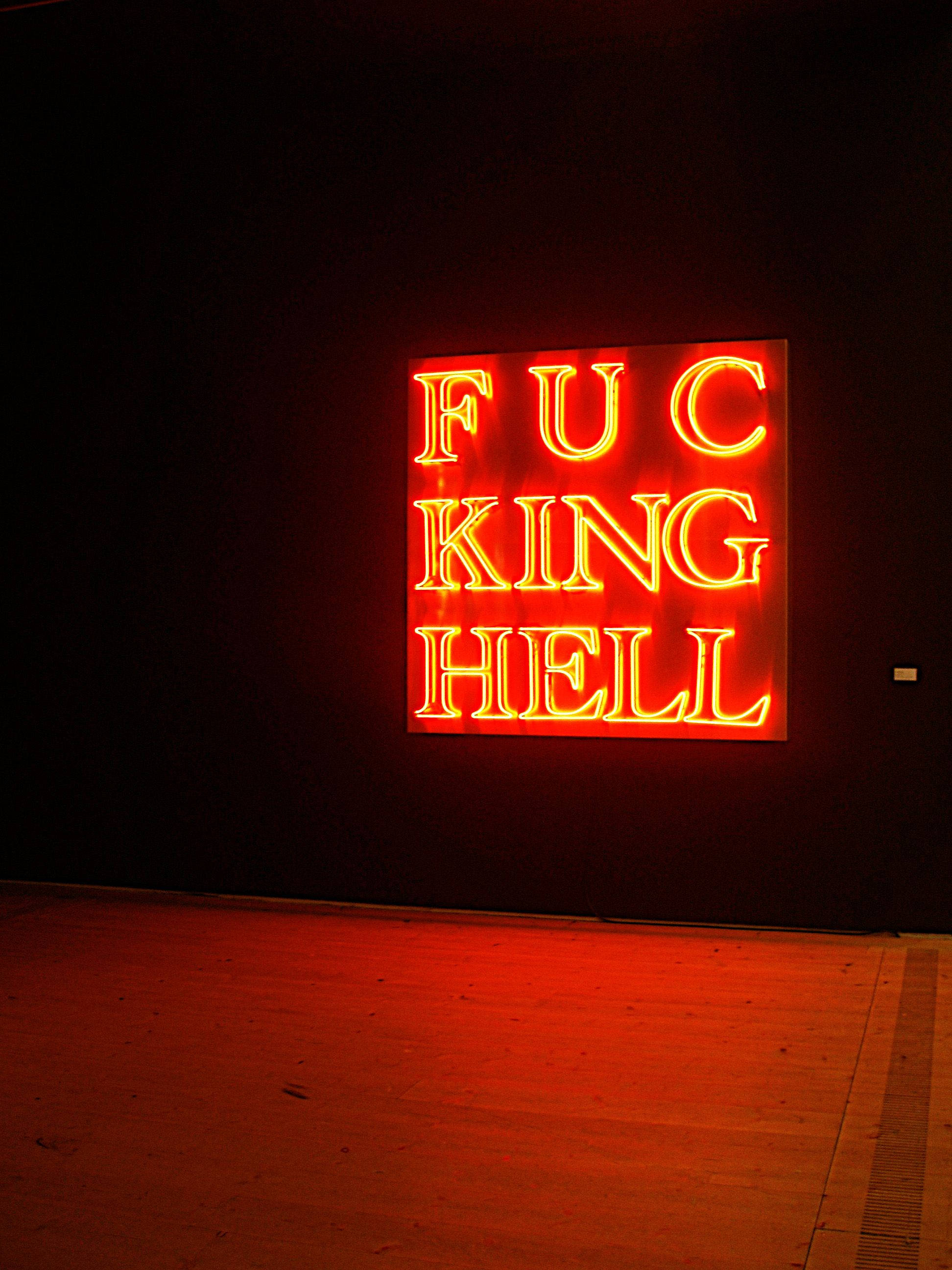 emo風格霓虹燈光 壁紙 — 一個發光的紅色霓虹燈牌，上面寫著「FUCKING HELL」，在黑暗的畫廊牆壁和木地板上投射出強烈的光線。
