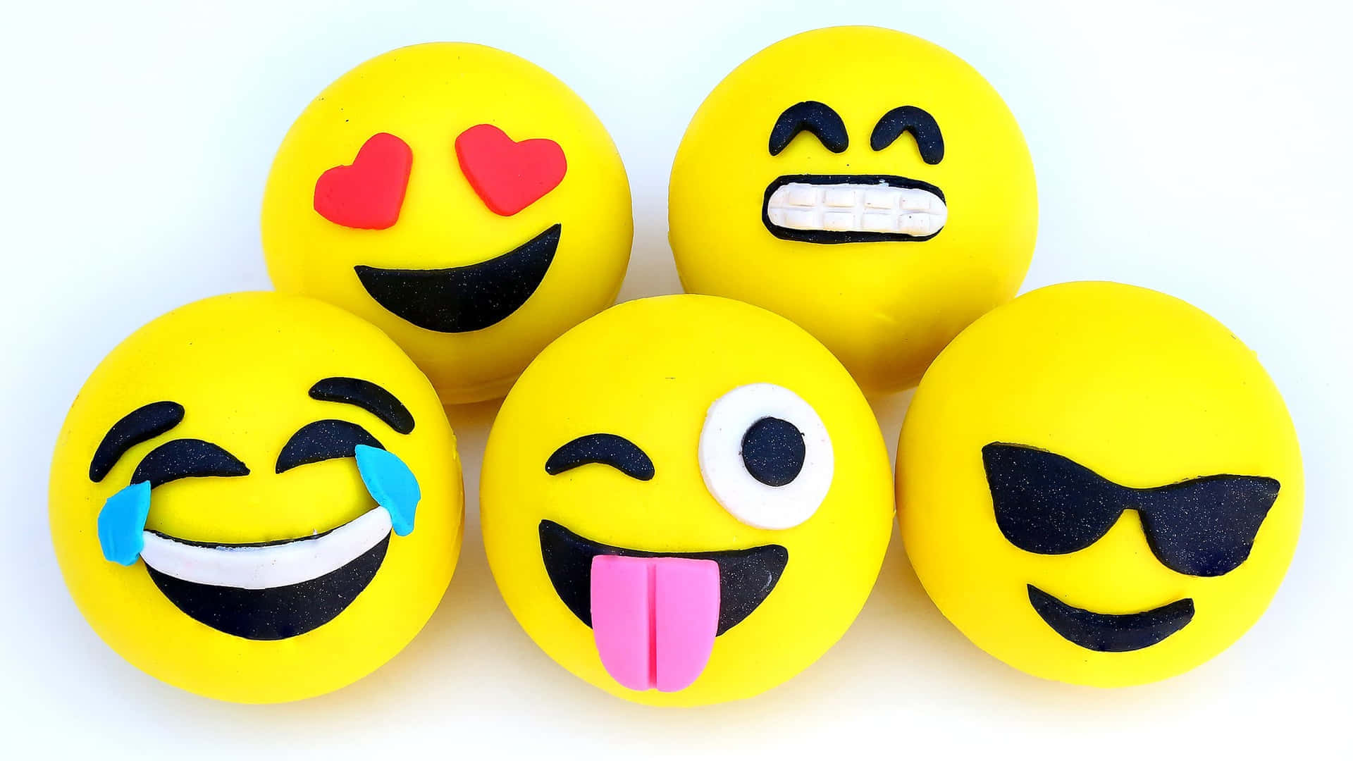 Emoji喜悅 壁紙 — 五個明亮的黃色表情符號公仔，帶有俏皮的表情，如心形眼睛、大笑吐舌頭和太陽眼鏡，坐在乾淨的白色背景前。