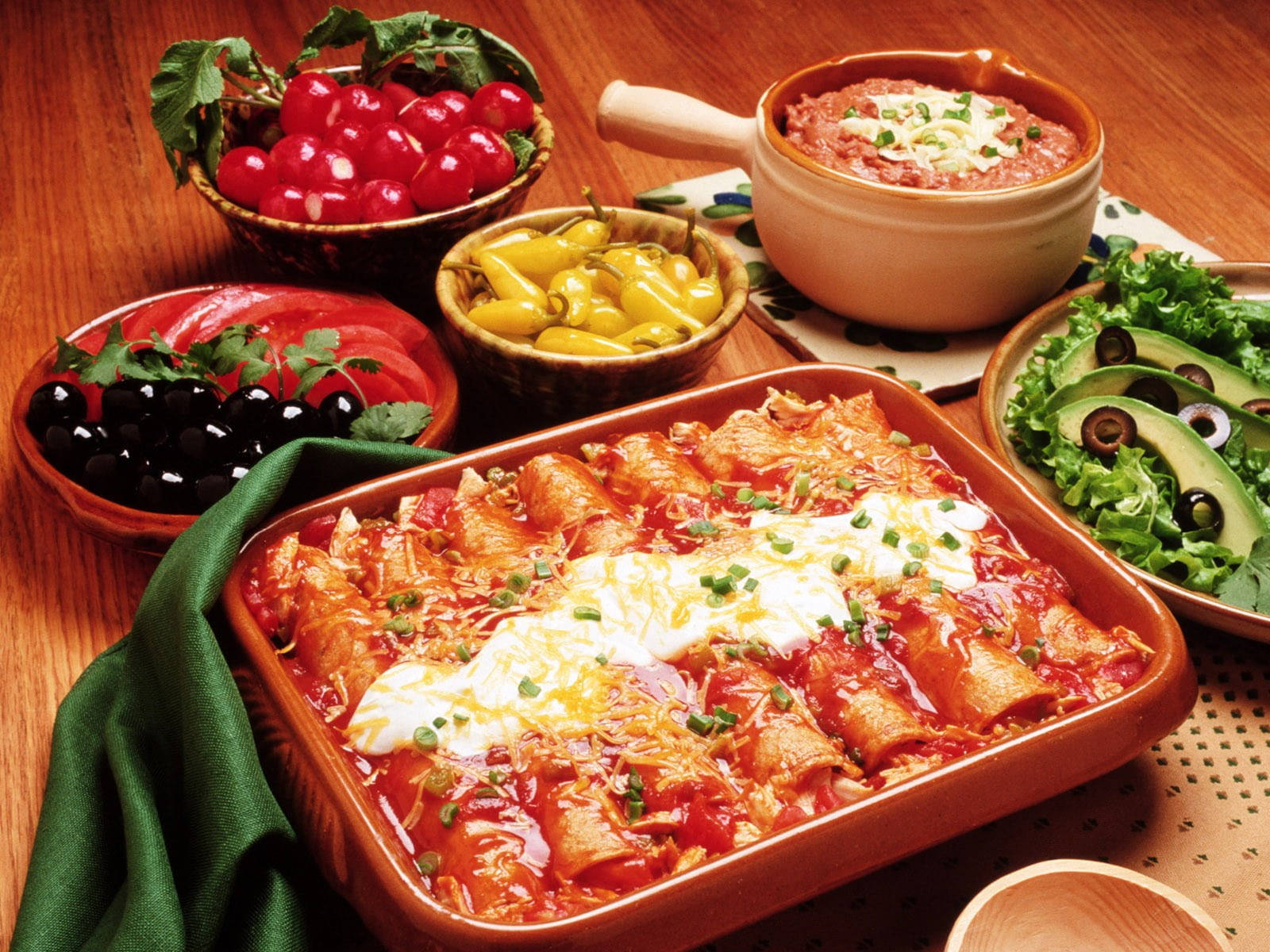 enchiladas 盤與生食材 壁紙 — 一頓充滿活力的墨西哥餐，有起司辣醬玉米餅配紅醬，周圍環繞著新鮮櫻桃番茄、橄欖和牛油果片，放在木桌上。