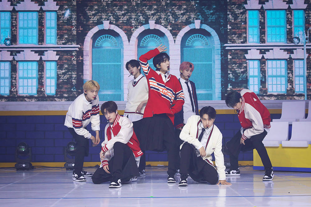 Enhypen《Tamed Dashed》舞台表演 壁紙 — 七位身穿紅、白、黑色棒球外套的男性K-pop偶像，在明亮的舞台上以磚砌建築為背景，擺出動感舞蹈姿勢。