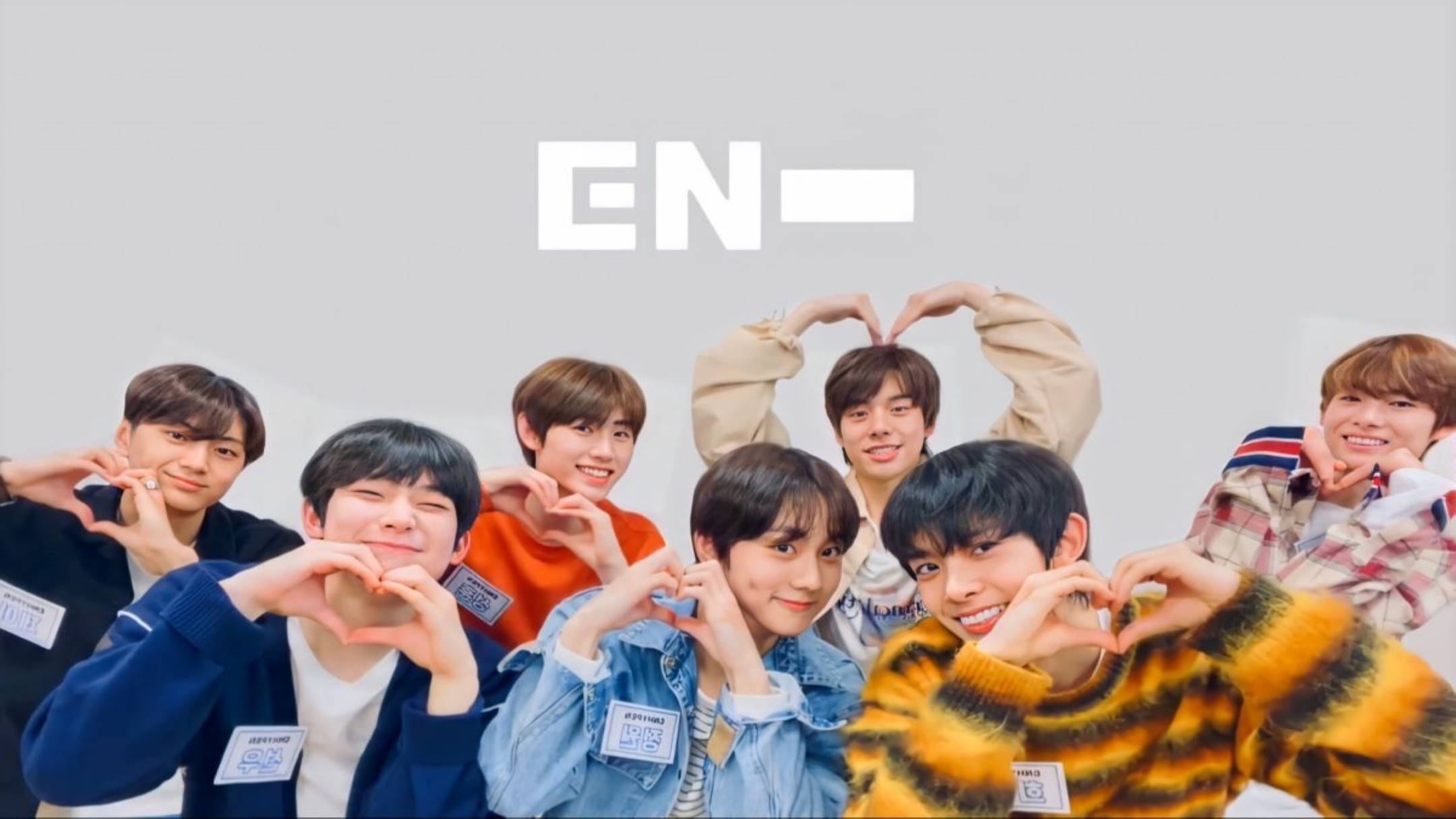 Enhypen V-Live現場 壁紙 — 七位微笑的年輕男士在純灰色背景前一起擺姿勢，穿著休閒服裝，用手做出心形。