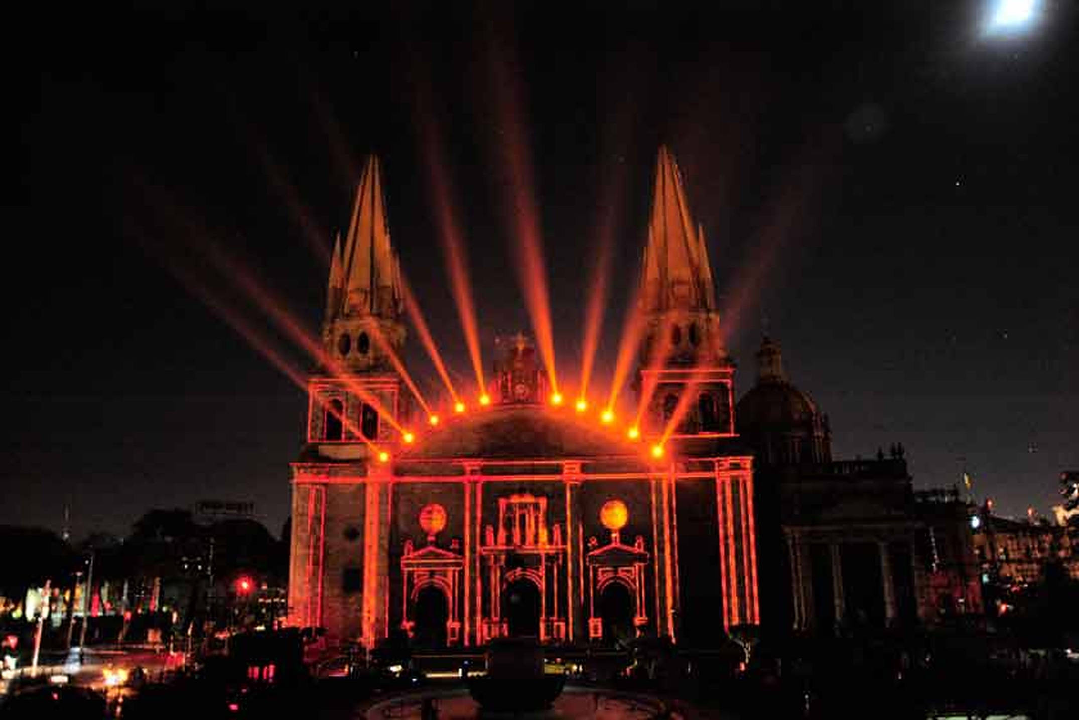 夜色中的 enlighten Guadalajara 大教堂 壁紙 — 一座宏偉的大教堂，被鮮豔的橙色激光束照亮，射向黑暗夜空，伴隨著明亮的月亮。