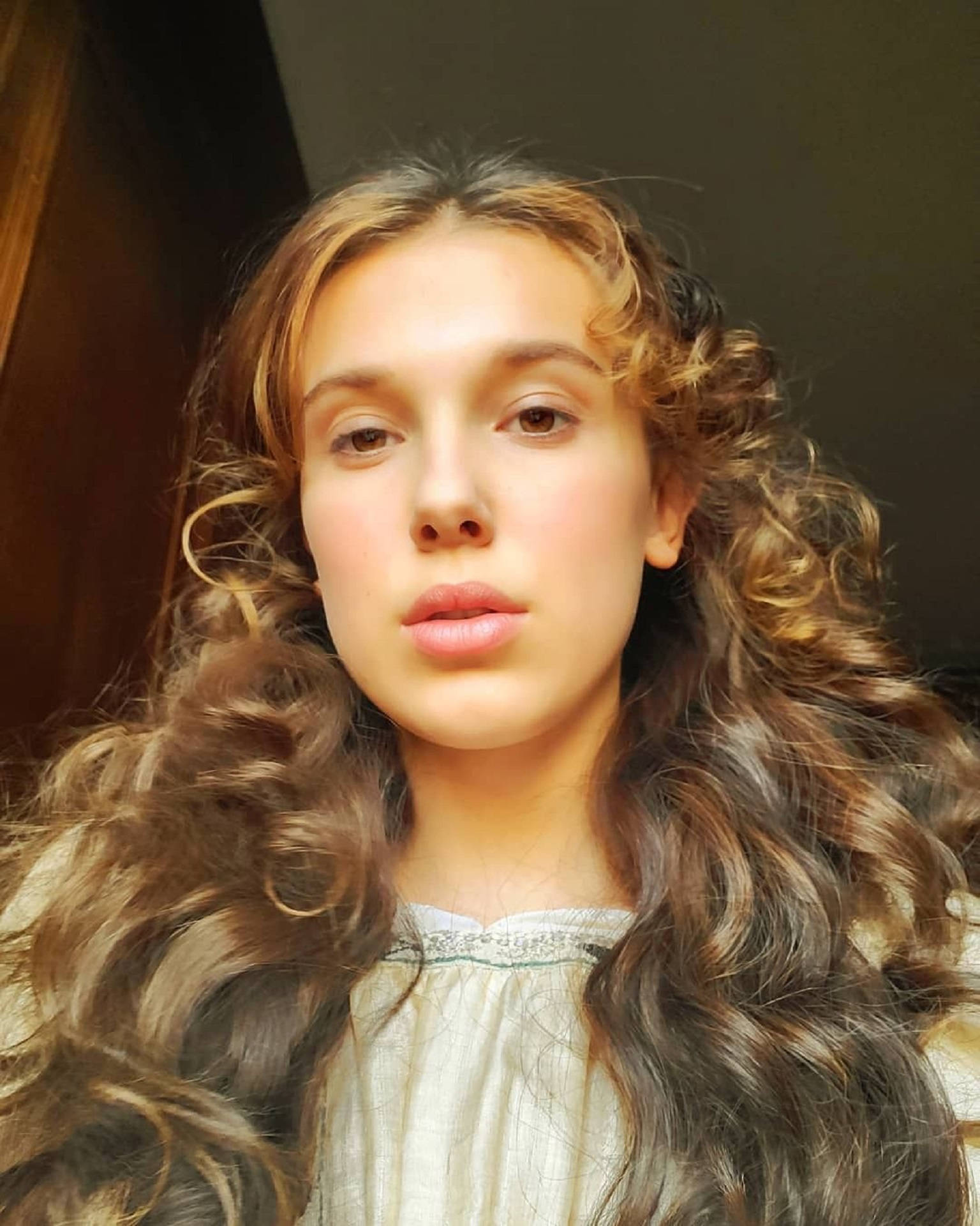 《Enola Holmes》米莉·波比·布朗Instagram長髮造型 壁紙 — 一位年輕女性的特寫肖像，擁有豐盈的棕色捲髮和柔和自然光線。