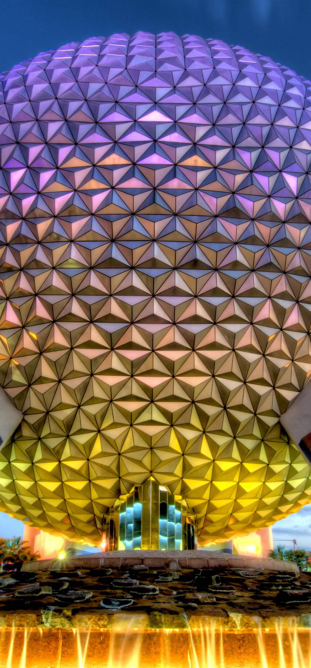 Epcot 球體與遊客 壁紙 — 一個巨大的測地線球體，帶有發光的紫色和金色三角形面板，在黃昏時聳立在噴泉上方。