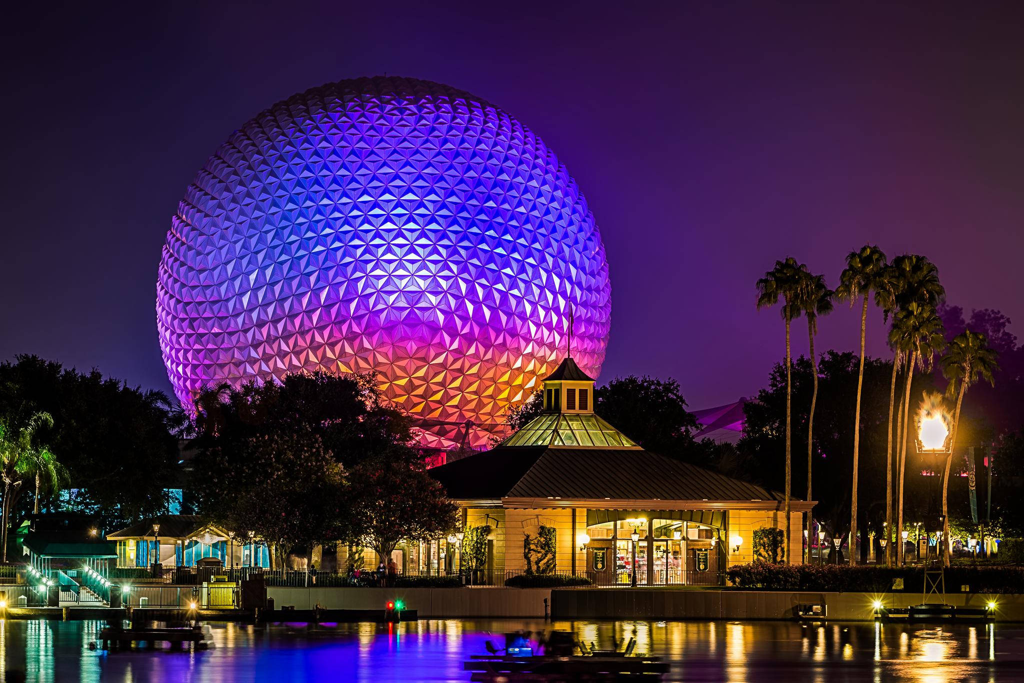 Epcot地球儀夜間發光 壁紙 — 一個巨大的測地線球體在夜間發出紫色和橙色的光芒，懸浮在海濱亭子上方。