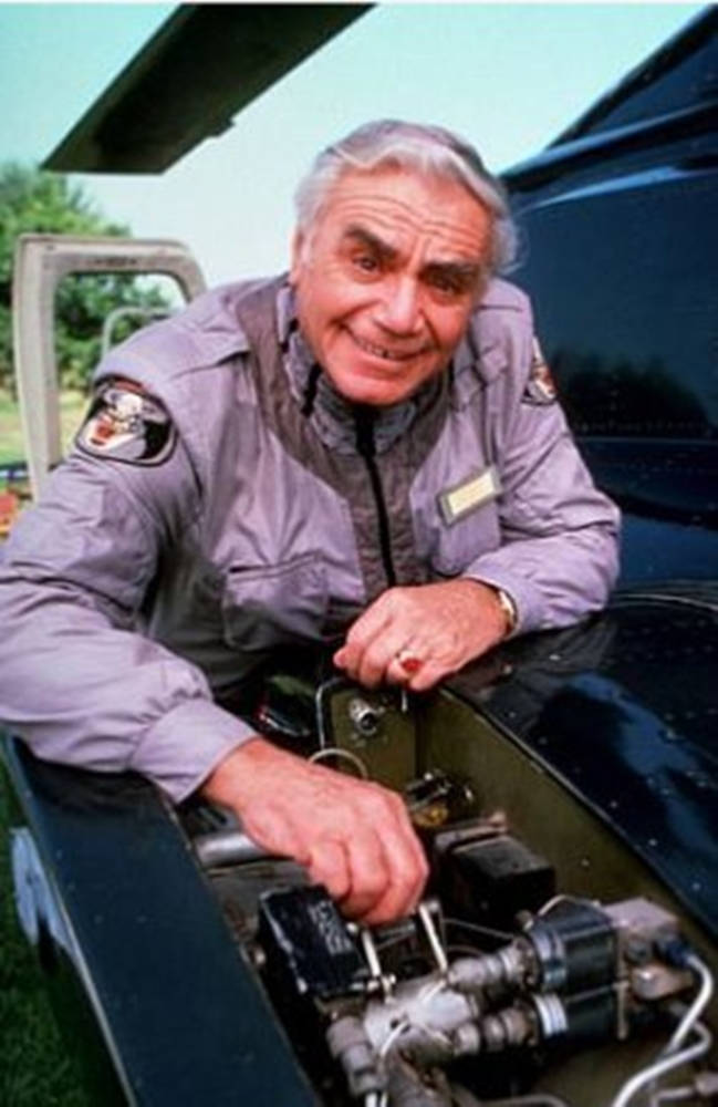 Ernest Borgnine 飾 Airwolf 中 Dominic Santini 壁紙 — 一位穿著飛行服的老年男子微笑著，正在打開引擎蓋的黑色飛機引擎上工作。