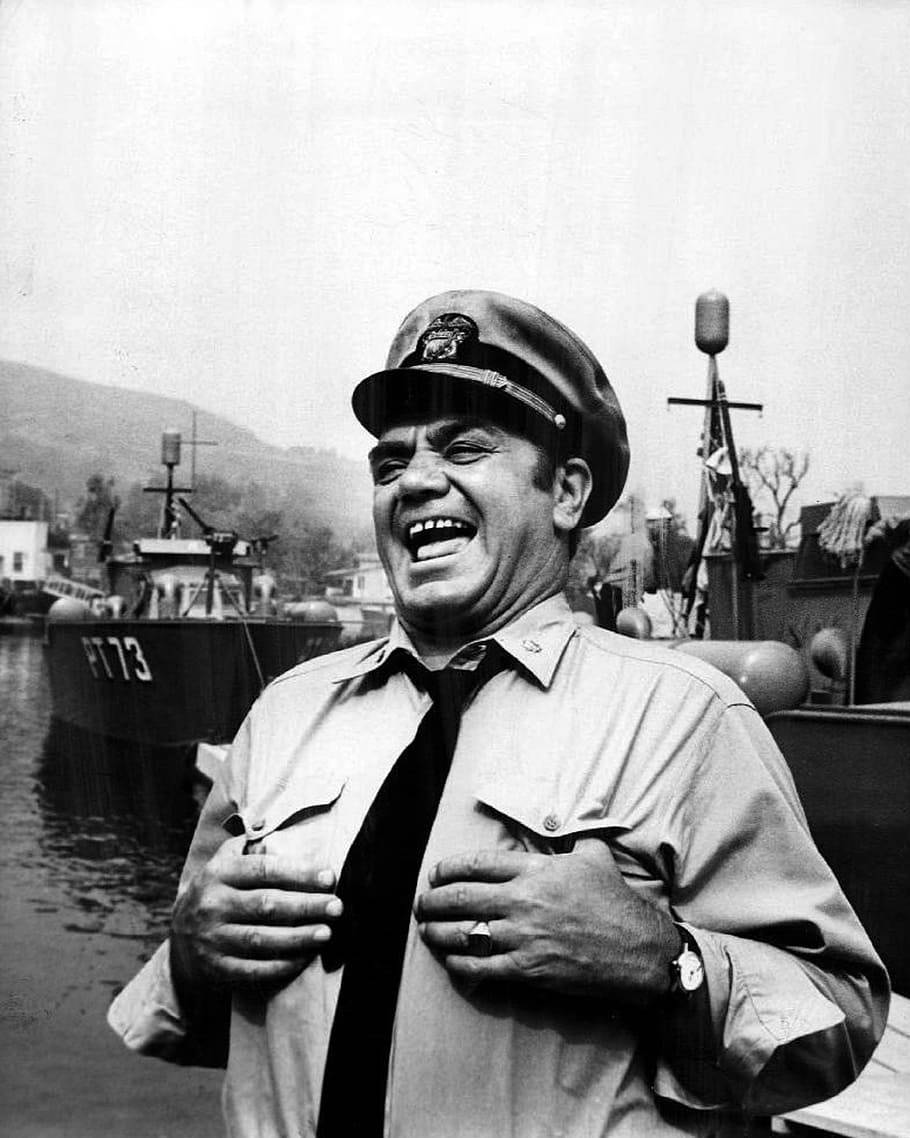 Ernest Borgnine 飾 McHale's Navy 中 Admiral Quinton McHale 壁紙 — 一名微笑的海軍軍官身穿制服，雙手放在胸前，在港口的一艘PT船旁開懷大笑。