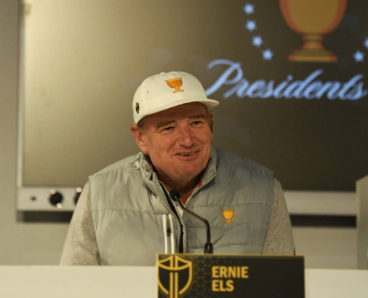 Ernie Els 壓力會議 壁紙 — 高爾夫球手厄尼·埃爾斯在記者會上微笑，戴著白色帽子及背心，上有金色獎盃標誌。