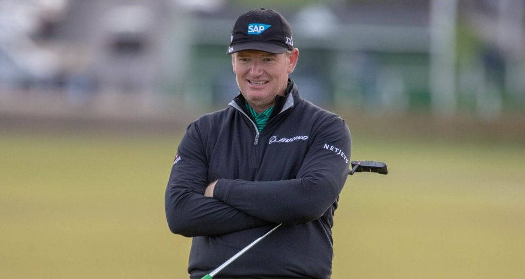 著名高爾夫球手 Ernie Els 跨臂擺姿 壁紙 — 一位戴黑色帽子和穿黑色外套的高爾夫球手微笑著，自信地交叉雙臂，在草地上握著推桿。